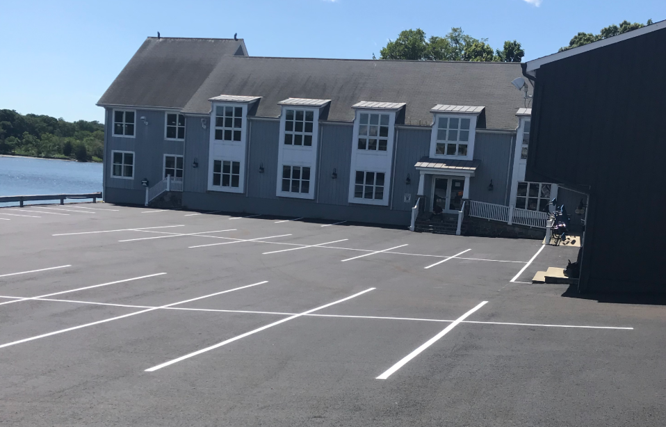 Empty parking lot in front of a blue building near a body of water, with some trees in the background.