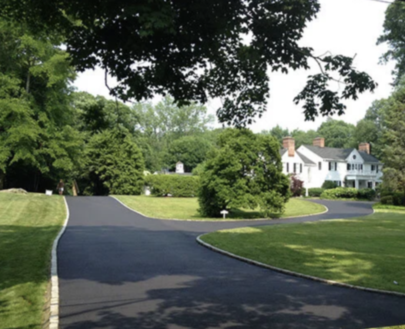 A winding asphalt driveway leading to a large white house with brick chimneys, surrounded by lush green trees and well-maintained lawn.