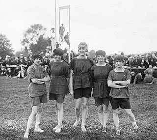 Five young girls in vintage athletic uniforms standing on a field, with a crowd and a high jump bar in the background.