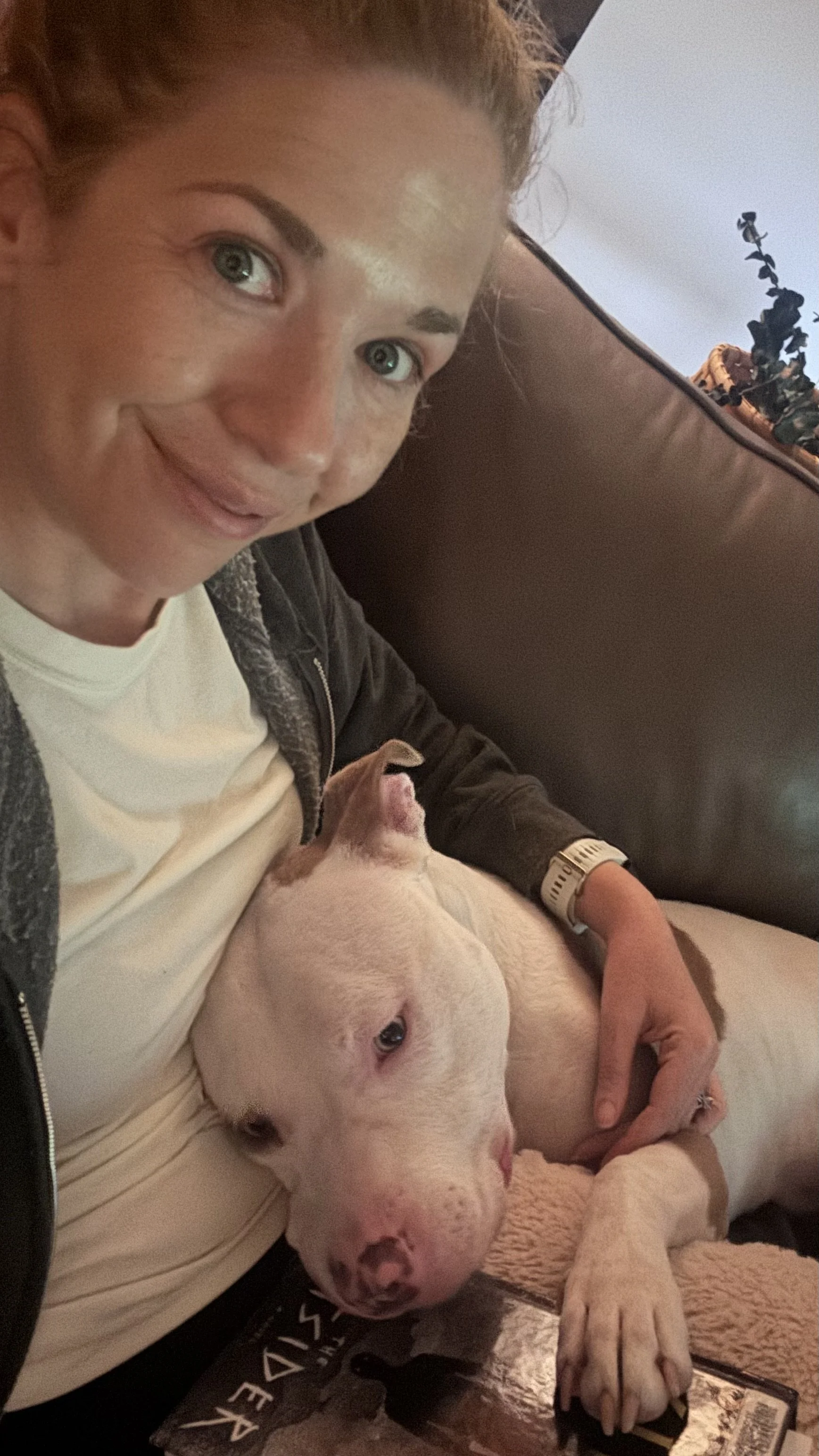 A woman with light skin and freckles smiling while holding a white puppy with black spots, lying on a blanket on her lap. She is sitting on a couch with a magazine nearby.
