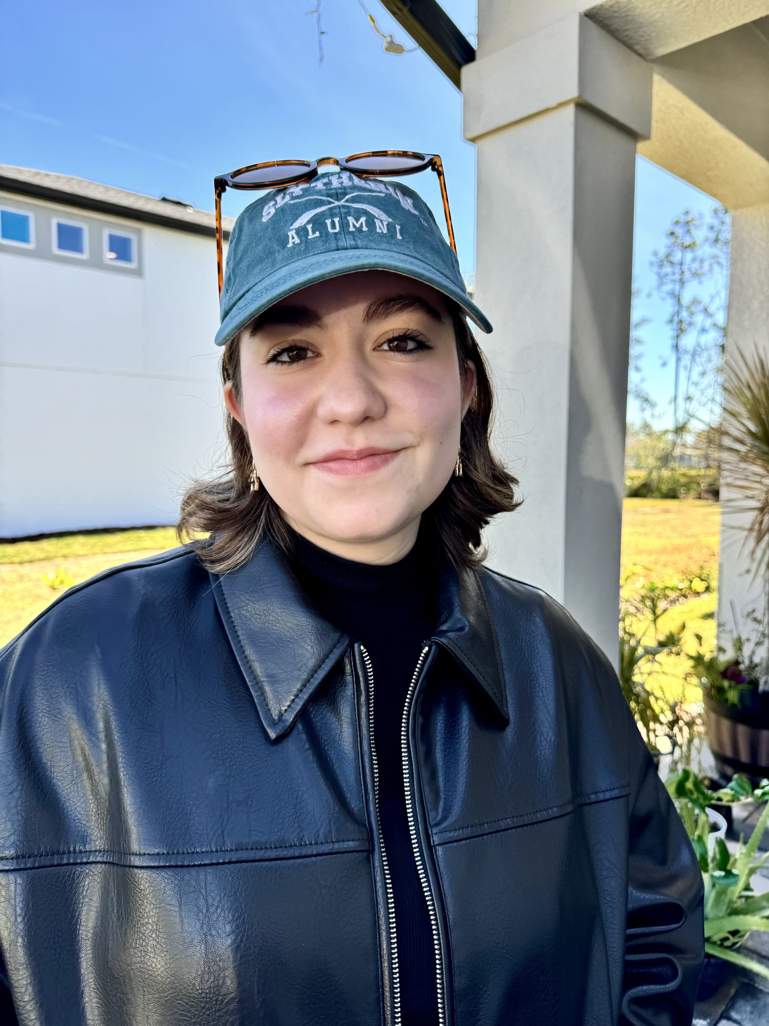A young woman with short brown hair, wearing a blue baseball cap with the words 'Senior Alumni' and sunglasses resting on top, is smiling at the camera. She is dressed in a black leather jacket and is outside in front of a modern building with plants and a bright blue sky in the background.