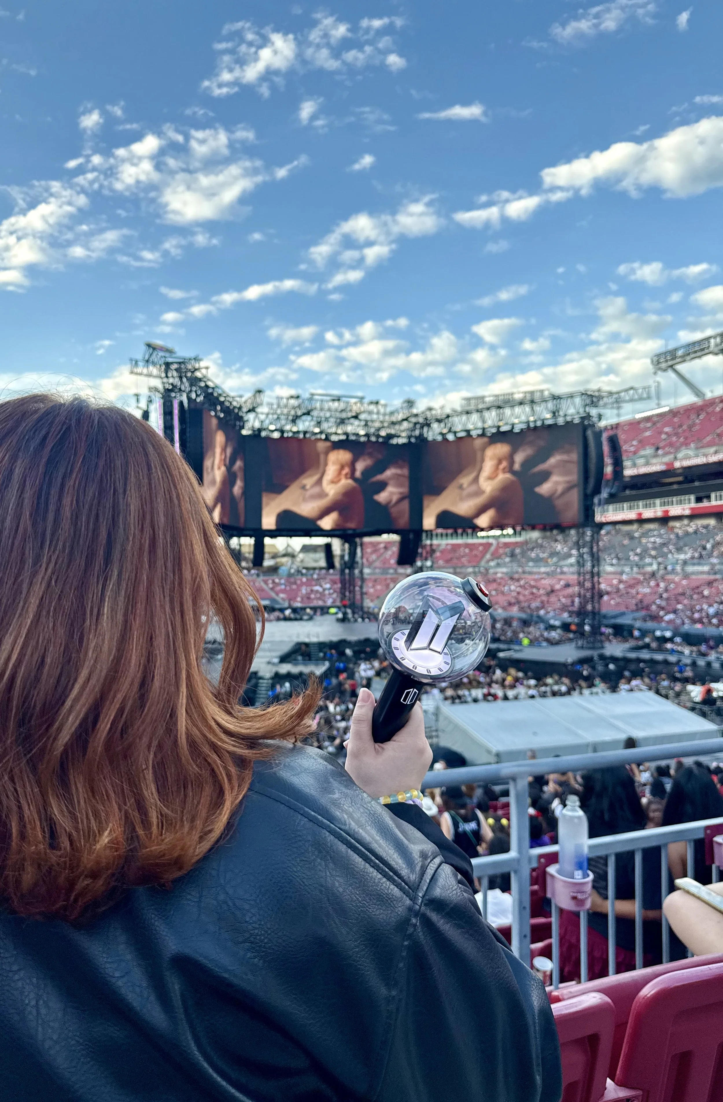 A person with reddish hair, wearing a black leather jacket, holding a fan with a silver handle and a clear plastic top, sitting in a stadium. In the background, a large outdoor concert stage with screens and an audience under a partly cloudy sky.