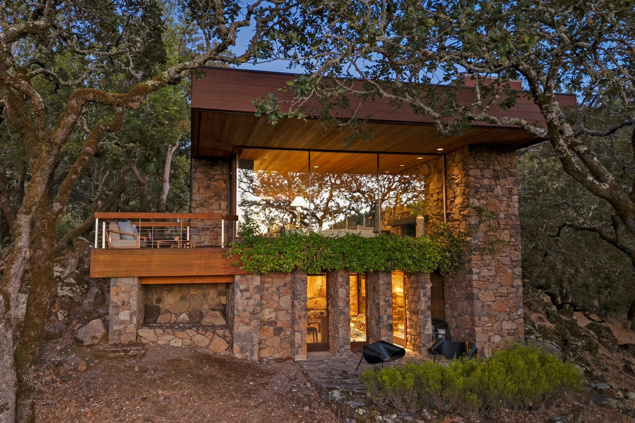 Modern house with large glass windows reflecting trees, stone accents, and a wooden balcony surrounded by trees at dusk.