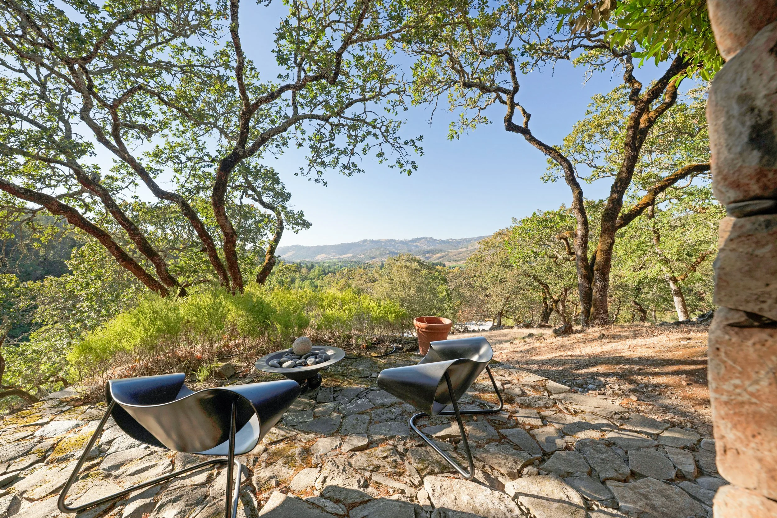 Outdoor patio with black chairs, a fire pit with rocks, a terracotta pot, and a stone paved ground under trees with green leaves. Hills are visible in the background.