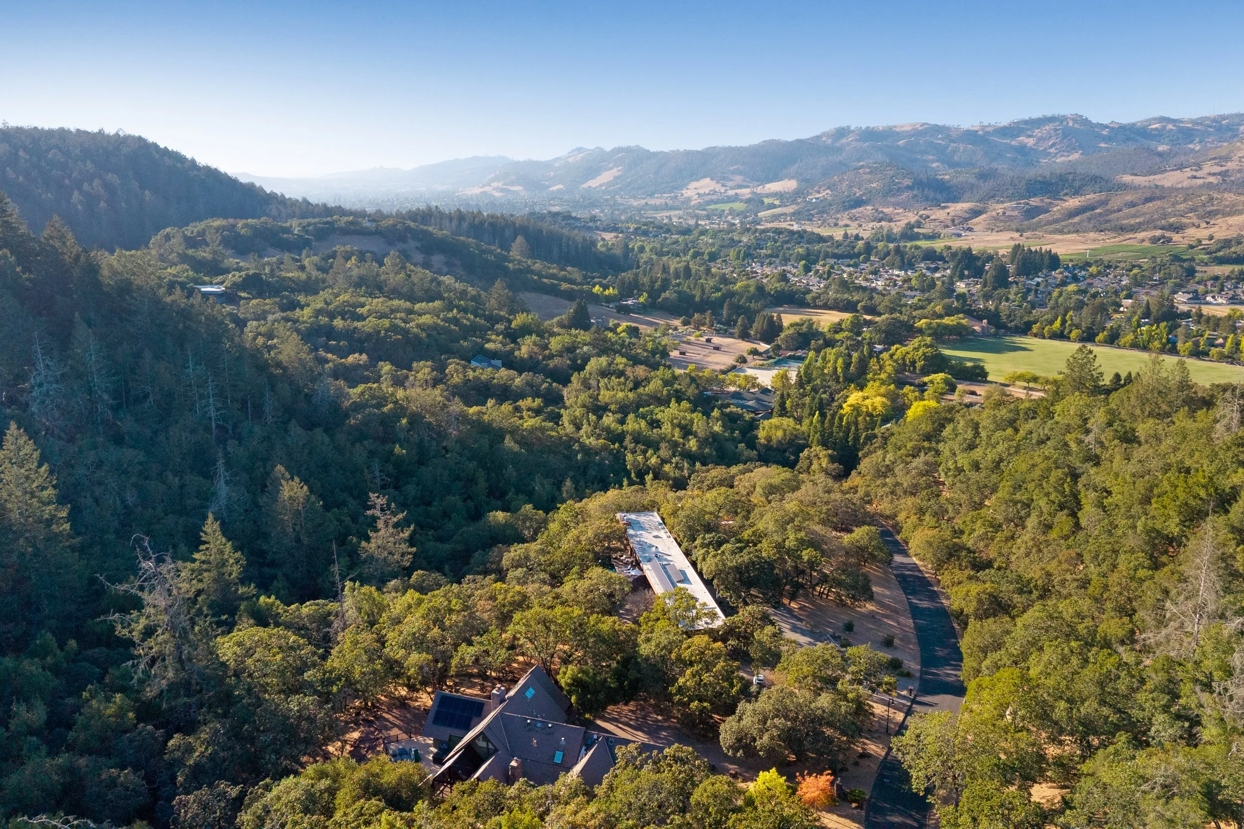 Aerial view of a green, hilly landscape with a small town and forested areas, mountains in the distance, and clear blue sky.