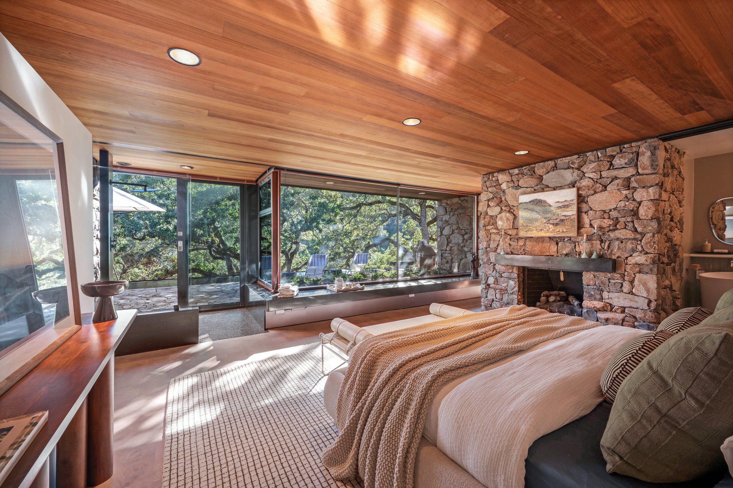A bedroom with a wooden ceiling and a stone fireplace. Large glass sliding doors lead to an outdoor area with trees and chairs. The bed is made with beige and striped bedding.