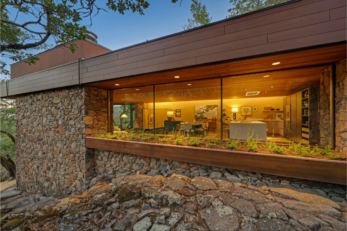 A modern house with stone walls and large glass windows, showing a cozy interior living room with furniture and lamps, situated on a rocky landscape surrounded by trees.