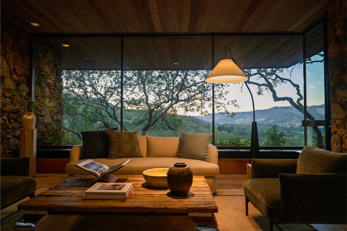 Living room with large windows showing trees and hills outside, a beige sofa with pillows, a coffee table with books and vases, a floor lamp, and other chairs.
