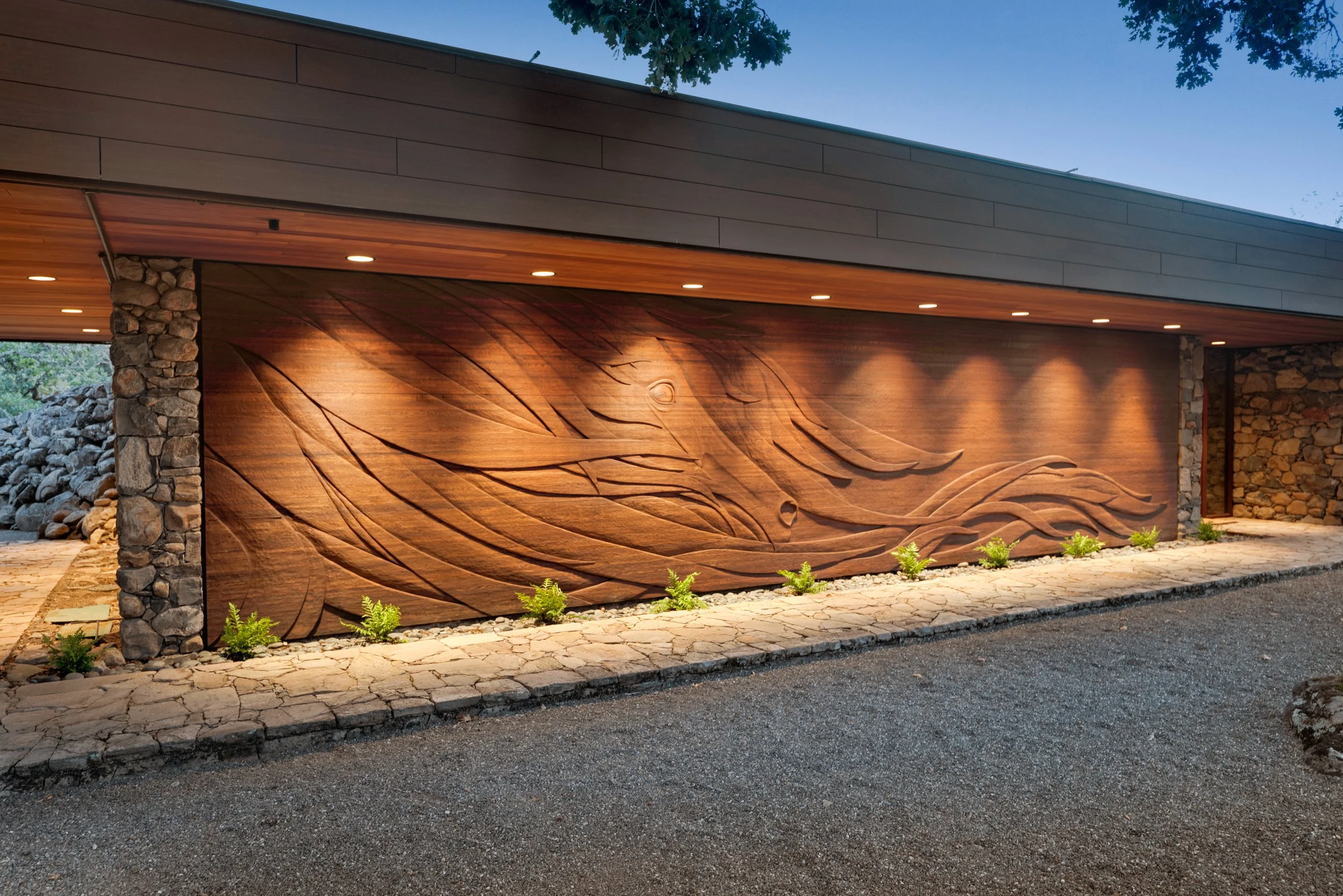 Modern building exterior with stone and wood wall design, small plants along the base, and a paved walkway in front. The roof has recessed lighting and the sky is visible with trees overhead.
