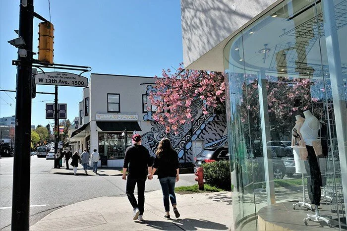 People walking on a sunny city sidewalk, with a mural on a building and a glass storefront with mannequins.