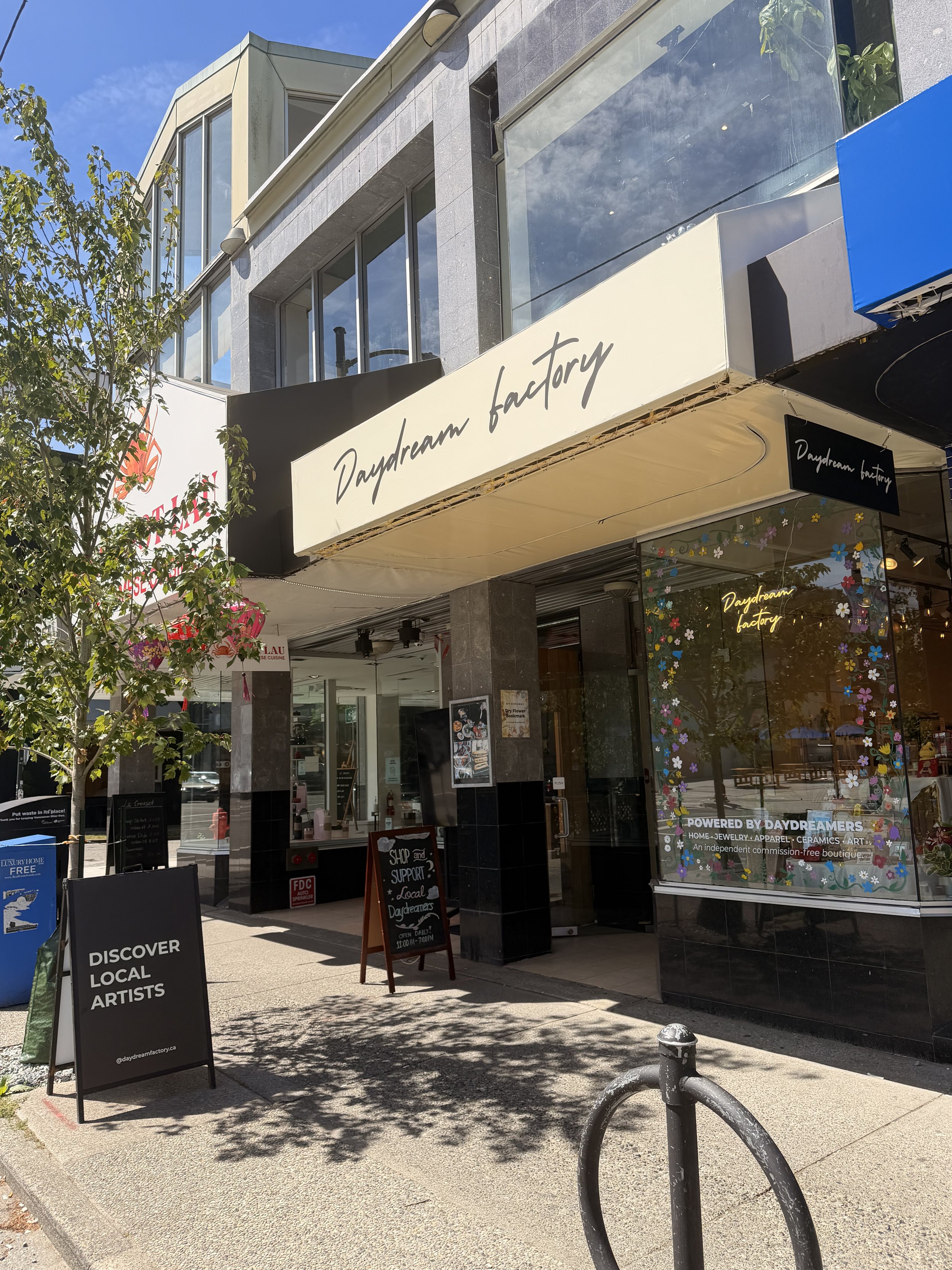 The exterior of a storefront named 'Daydream Factory' with signs encouraging supporting local artists and shopping local. There are trees, benches, and a sidewalk in front of the shop, with a blue sky above.