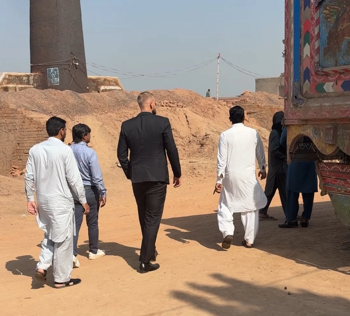 A group of five men walking across a dry, sandy area in a desert-like setting. Some are dressed in traditional white clothing, one in a black suit, and others in casual attire. There is a large, painted truck on the right side and a tall mud-brick structure in the background.