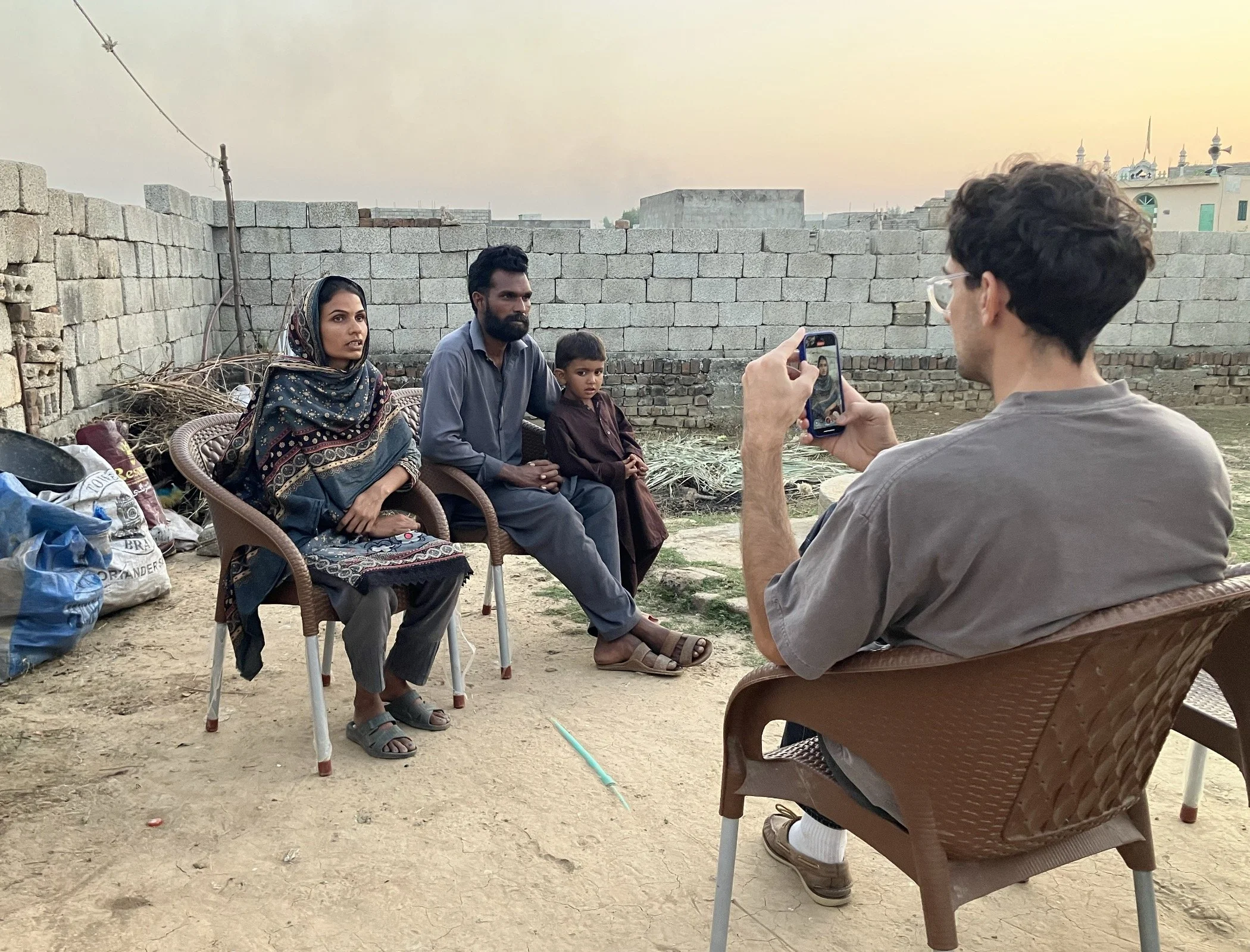 A man with glasses and curly hair taking a photo of a family sitting on chairs against a brick wall in an outdoor setting at dusk. The family includes a woman wearing a headscarf, a man with a beard, and a young boy. Bags and debris are visible on the ground.