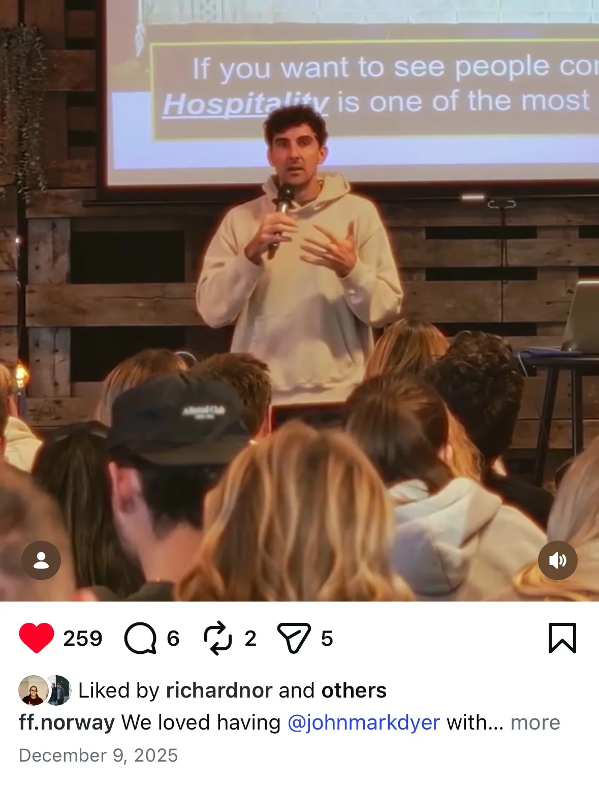 A man holding a microphone giving a presentation in front of a screen with text, while an audience listens. The setting appears to be a rustic indoor space with wooden walls.