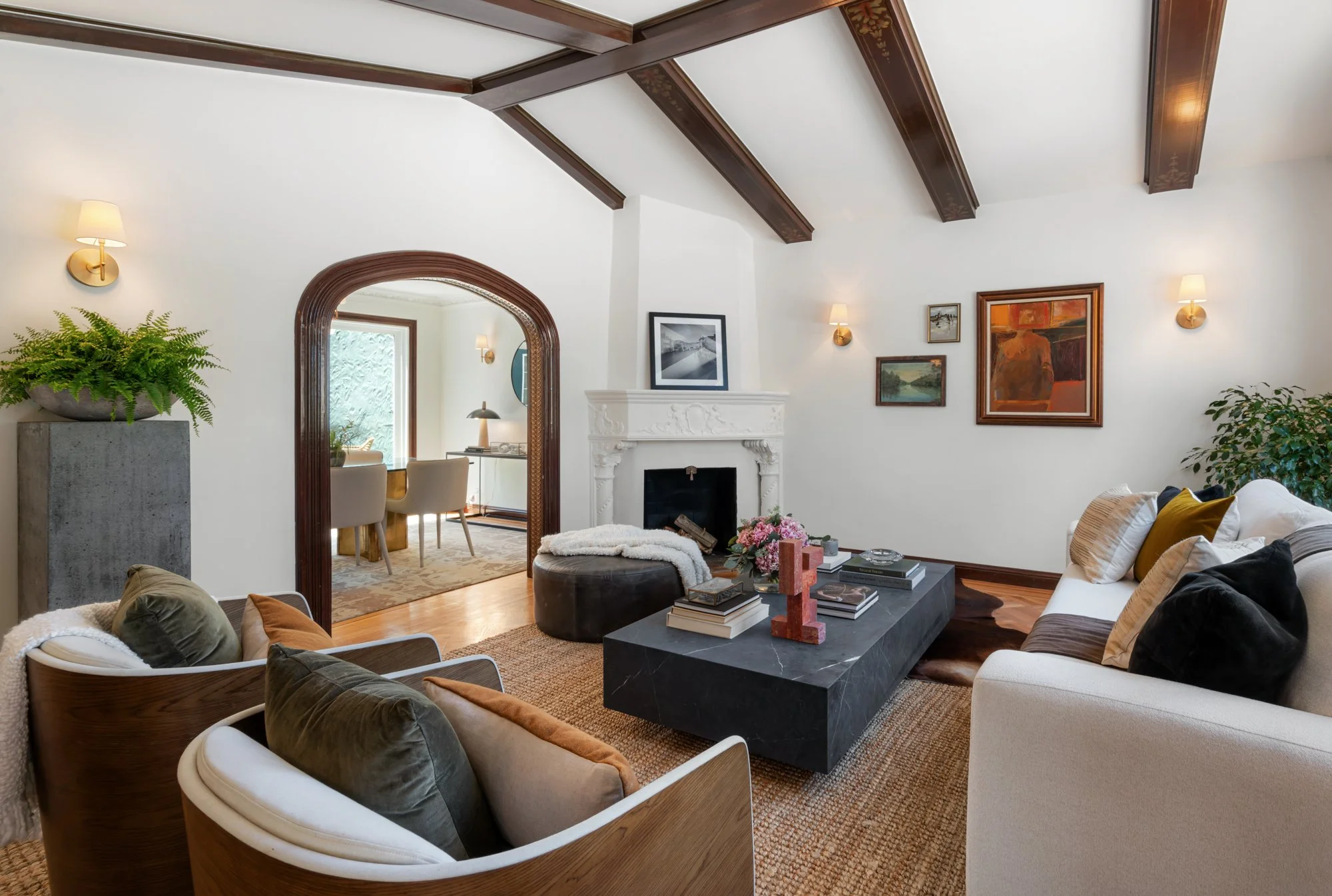 Living room with wood-beamed ceiling, white walls, various framed paintings, a fireplace, and modern furniture including a white couch, cowhide ottoman, and black marble coffee table with decorative items
