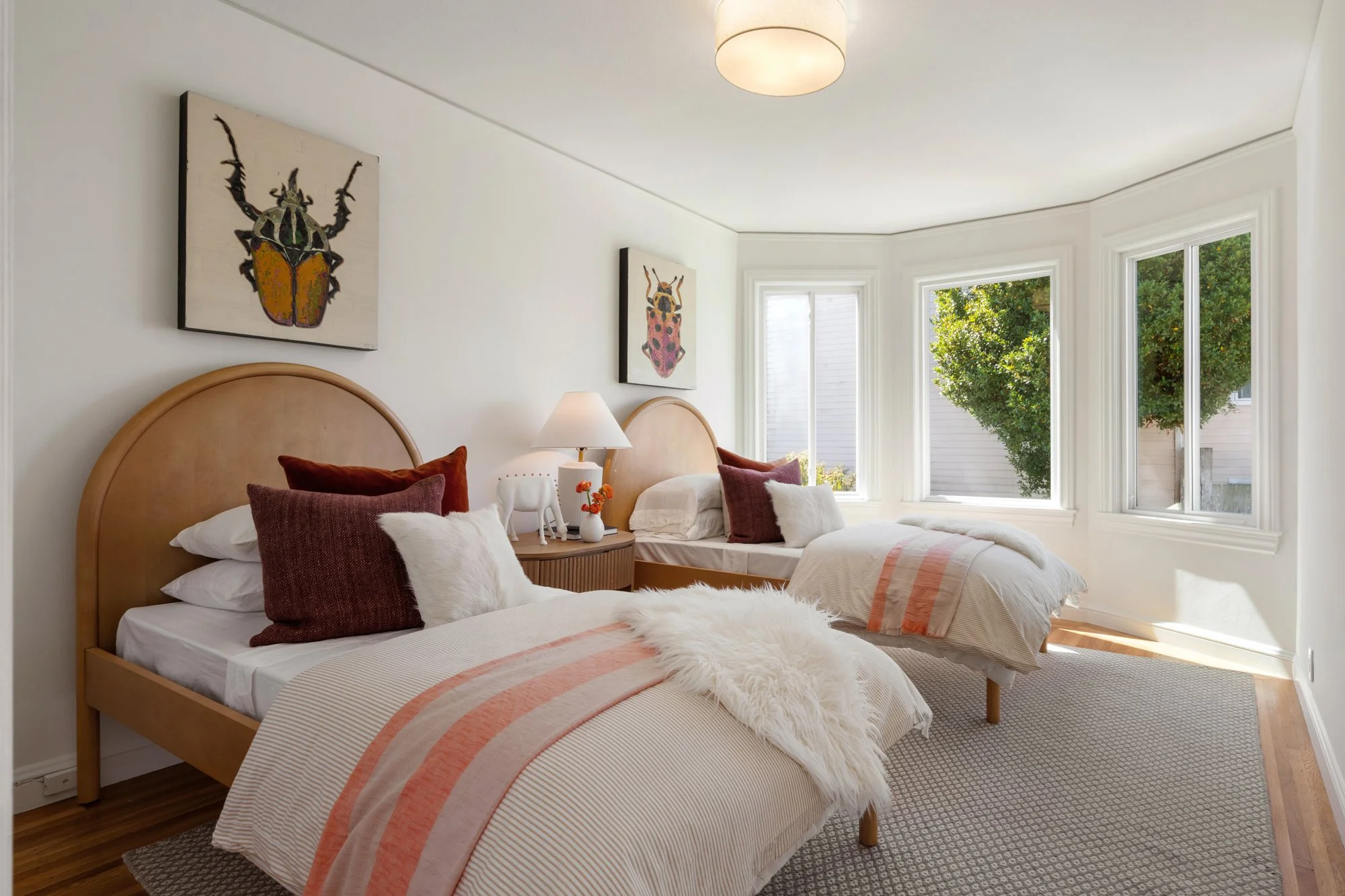 A bright bedroom with two wooden beds, each with beige and burgundy pillows, white and fluffy throws, and a striped beige and orange bedspread. A wooden nightstand between the beds holds a white lamp, small decorative items, and a ceramic elephant. T