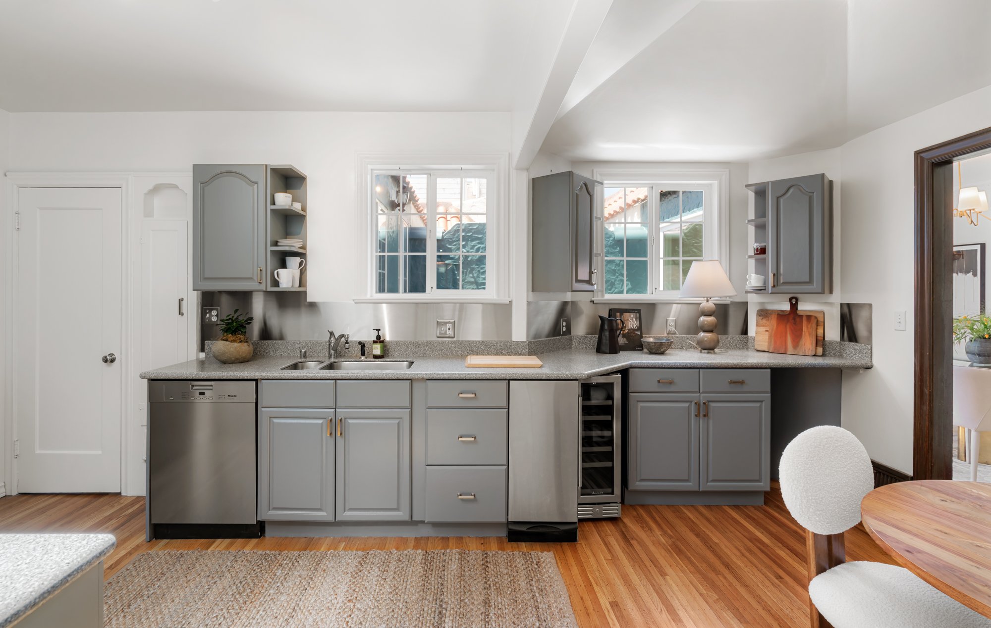 Modern kitchen with grey cabinets, a speckled grey countertop, and two windows providing natural light. The kitchen features a built-in sink, dishwasher, wine fridge, and open shelves with dishes and a plant. Part of a round dining table and an uphol