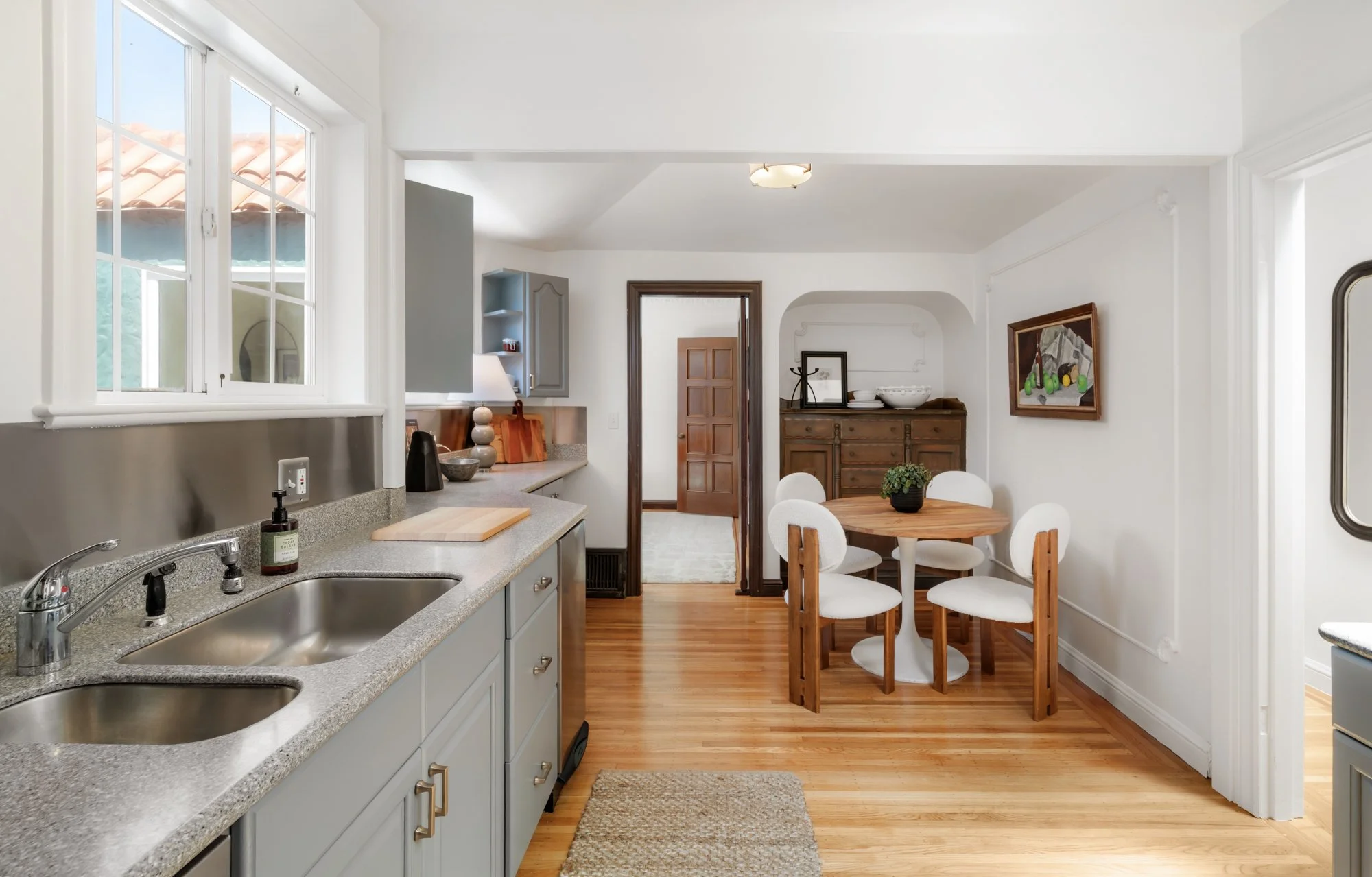 A kitchen with white cabinets, a gray countertop, a double sink, and a wooden dining table with white chairs. There is a wooden sideboard with decorative bowls and framed pictures. Large windows let in natural light, and hardwood flooring is visible.