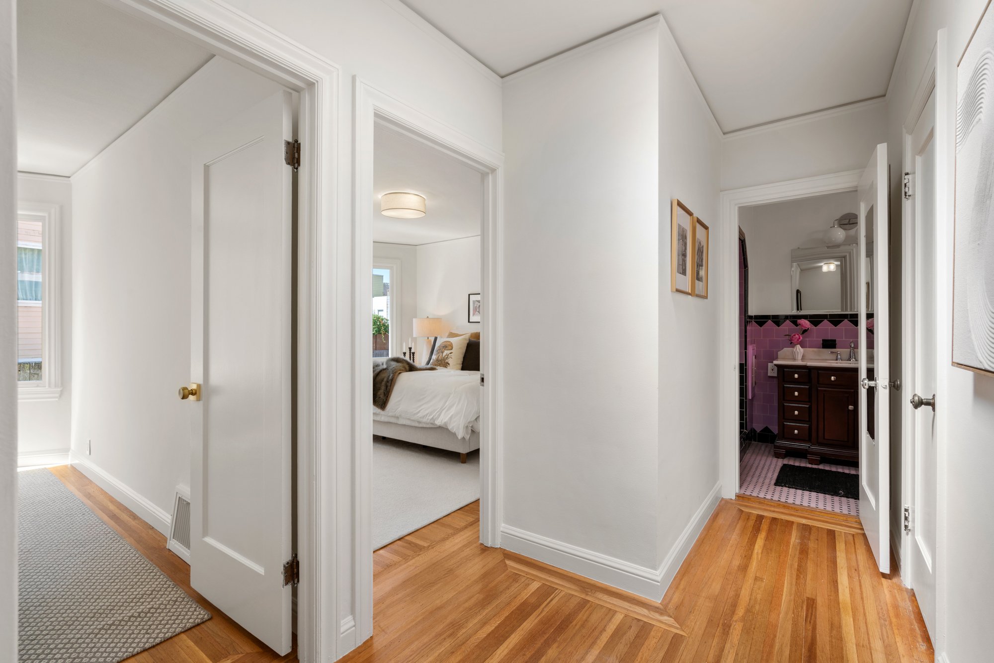 A hallway with white walls and hardwood floors, leading to a bedroom with a bed, lamp, and framed pictures, and a bathroom with purple tiles and a dark wood vanity.