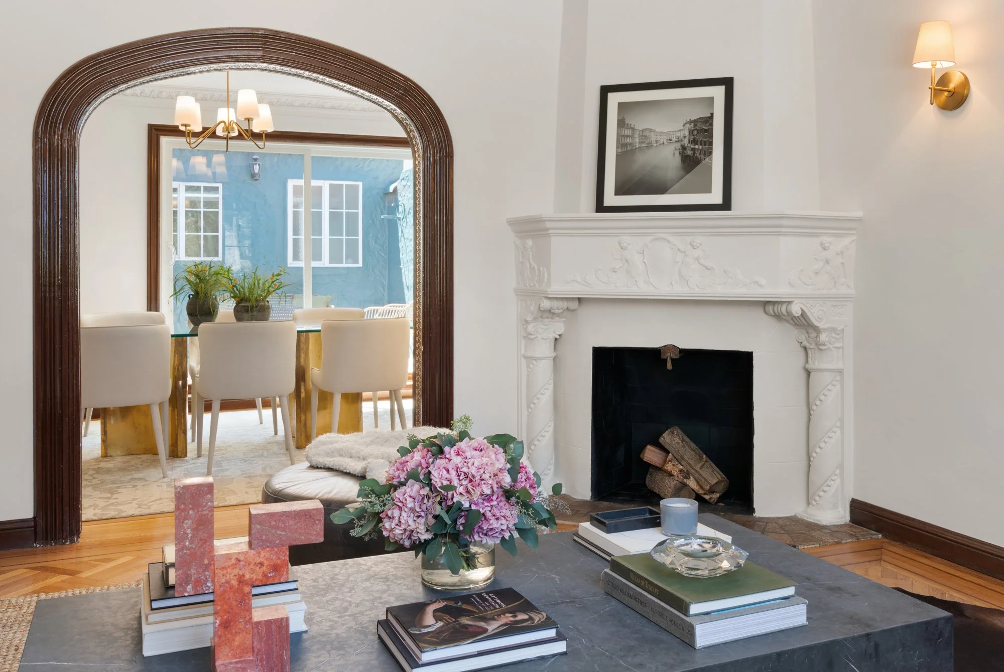Living room with white walls, a fireplace with ornate white mantel, black-and-white photograph above it, and a wall-mounted sconce light. In the foreground, a table features pink hydrangea flowers, art books, and pink granite samples. Through an arch