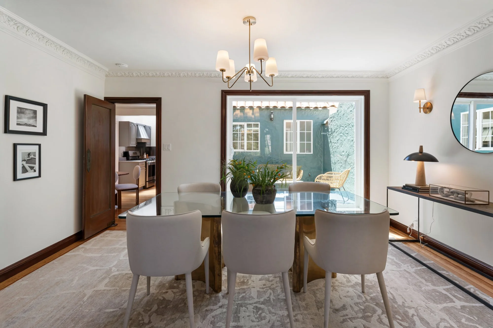 Dining room with a glass-top table, six white chairs, and large windows showing an outdoor patio with chairs. Light fixtures include a chandelier and wall sconces, with framed photos on the wall and a sideboard with objects and a round mirror.