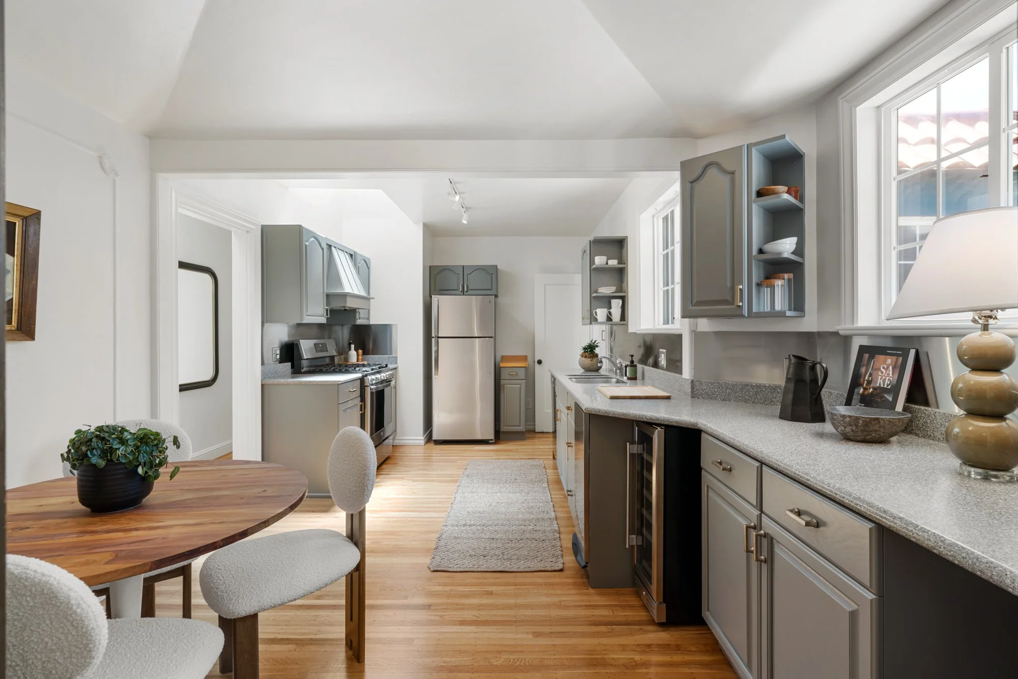 Modern kitchen with gray cabinets, stainless steel refrigerator, stove, and beige countertops. Includes a circular wooden dining table with white upholstered chairs, a potted plant, and a window with natural light.