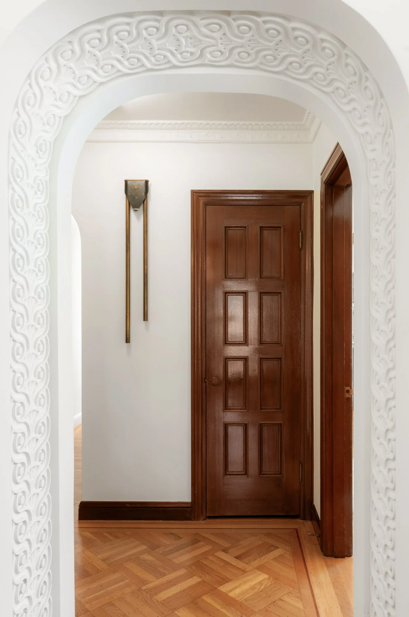 Interior view of a hallway with a wooden door, hardwood flooring, decorative white arch with intricate patterns, and a wall-mounted brass and gray umbrella stand.