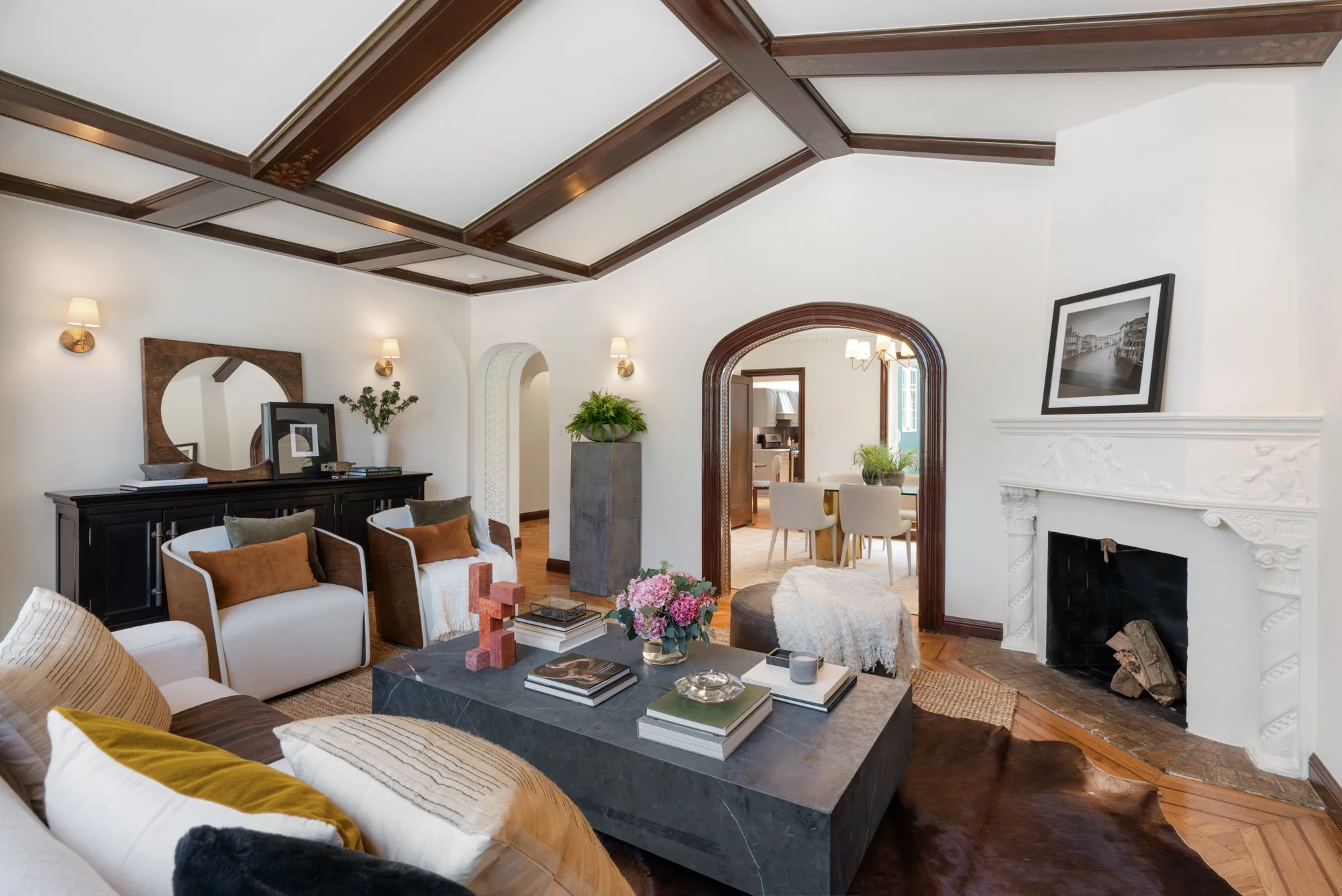 Living room with white walls, wood ceiling beams, and a fireplace. Furniture includes a white sofa with yellow and black pillows, armchairs, a coffee table with decorative objects, and a sideboard. There are wall-mounted lamps, framed black-and-white