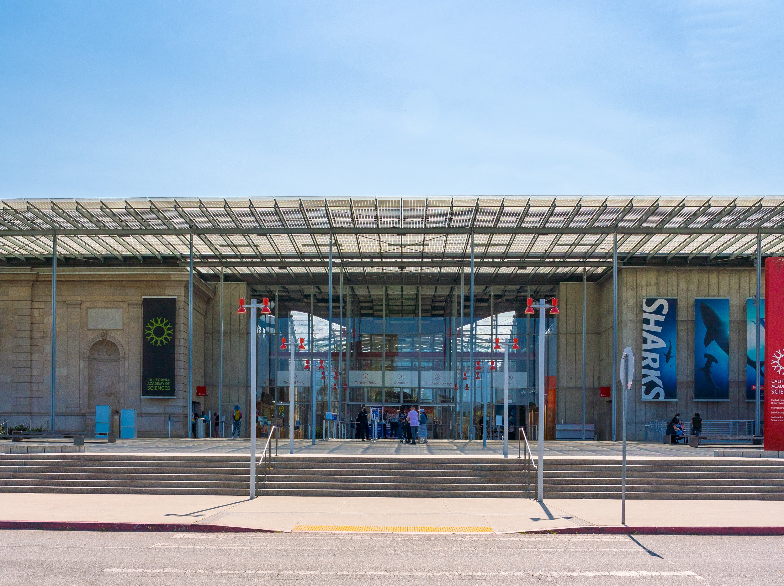 Entrance to a building with steps and a large glass roof, people walking in and out, banners on sides, and flags and signs at front.