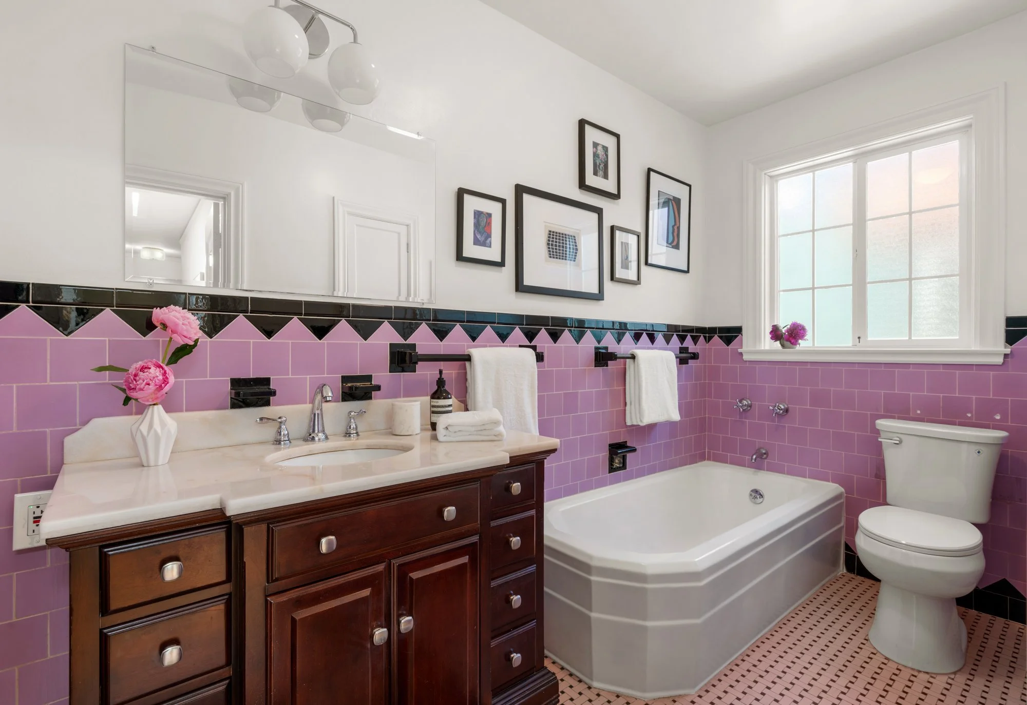 Bathroom with pink tile backsplash, white walls, a large window, and dark wood vanity with marble countertop. Pink flowers in a white vase on the sink, framed artwork on the wall, towel racks with white towels, and a bathtub with fixtures.