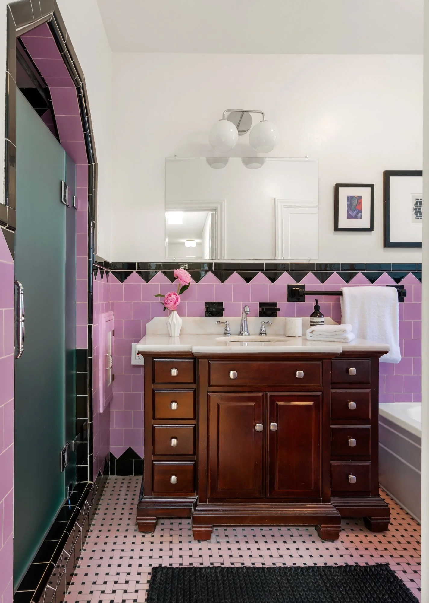 A vintage-style bathroom with a pink tiled wall, dark wood vanity with marble countertop, a large mirror, and framed artwork. Pink flowers in a white vase sit on the counter, and a black-and-white patterned rug is on the black-and-white tiled floor.