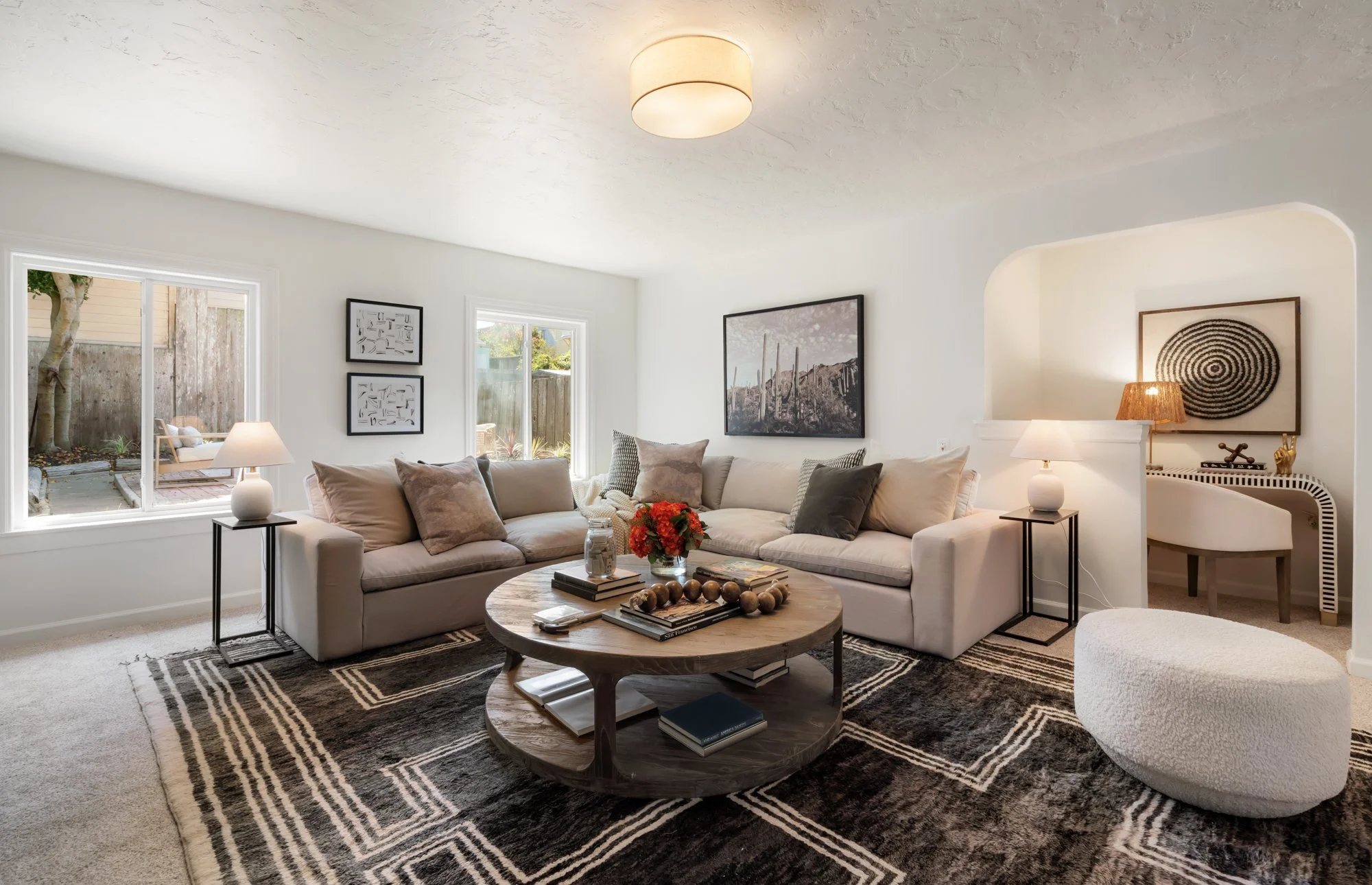 A living room with a white L-shaped sofa adorned with several gray and beige pillows, a round wooden coffee table with books and decorative objects, a black and white patterned area rug, and artwork on the walls. Large windows let in natural light, a