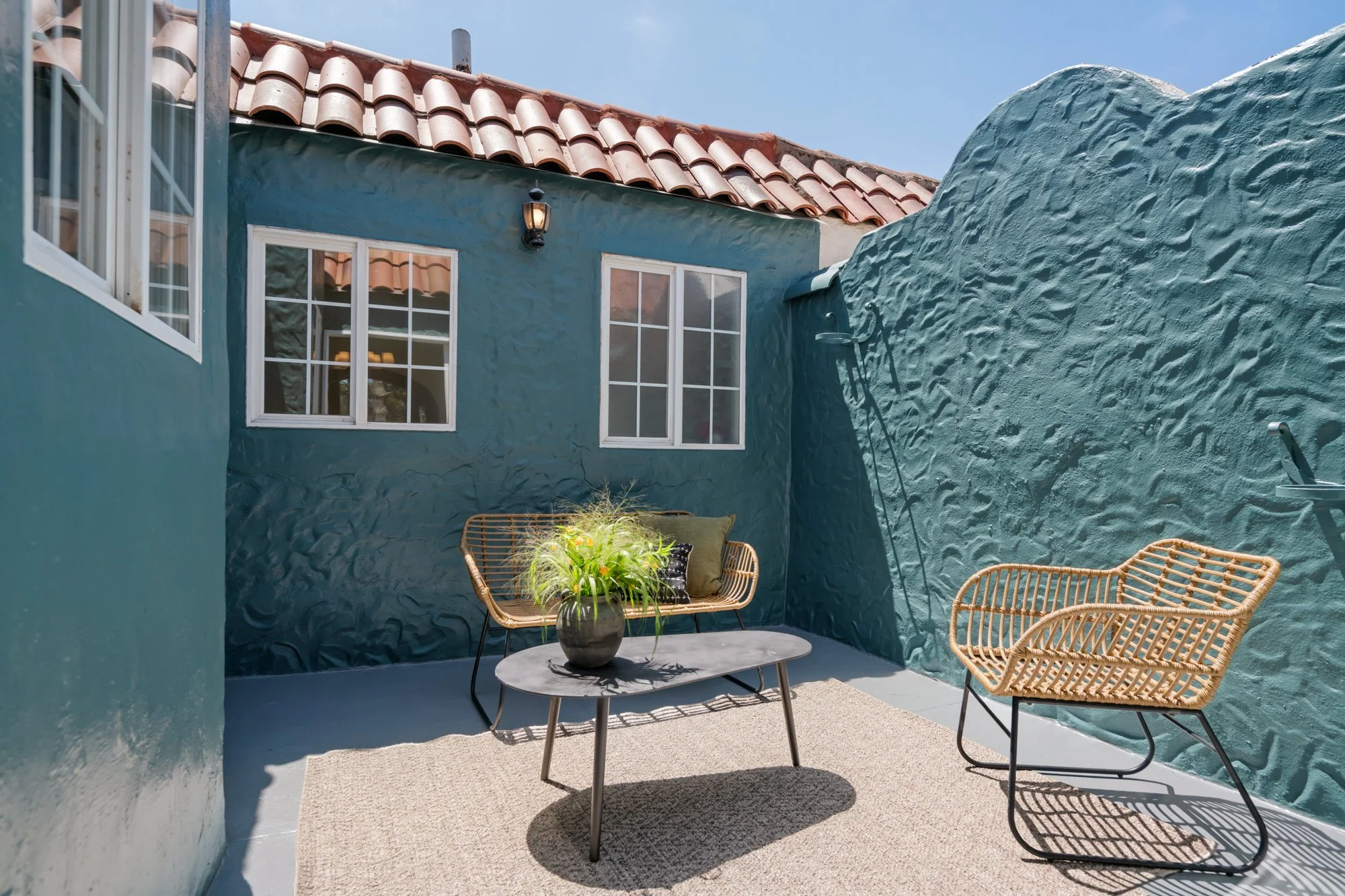 Patio with a round table, potted plant, wicker chairs, a blue stucco wall, windows, and a tile roof, under a clear blue sky.