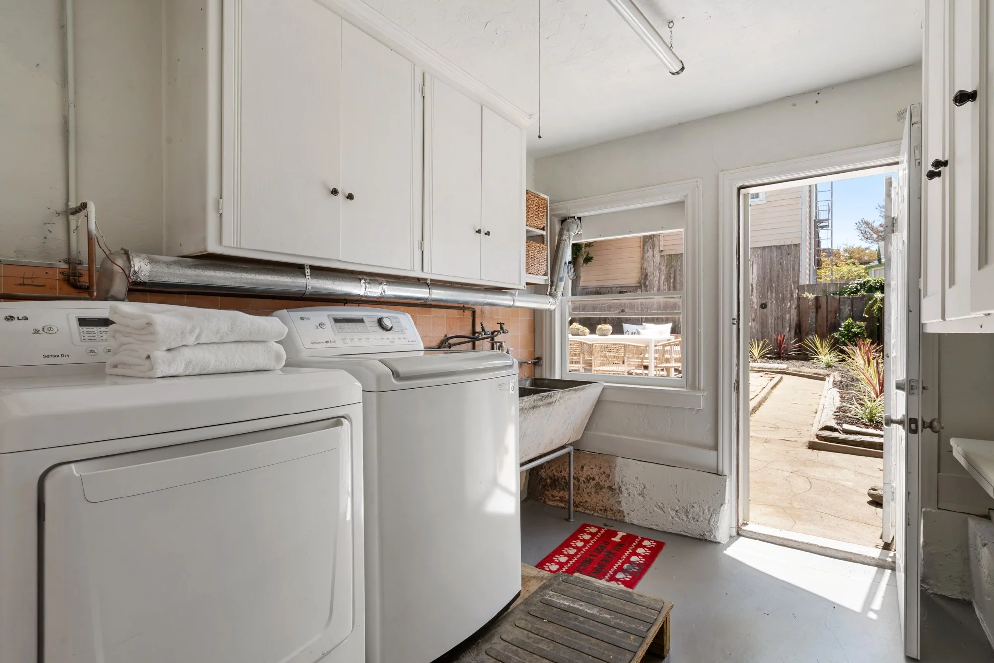 A small laundry room with white washing machine and dryer, white cabinets above, laundry towels on the washing machine, a utility sink, and a door leading outside to a patio with outdoor seating, plants, and a wooden fence.