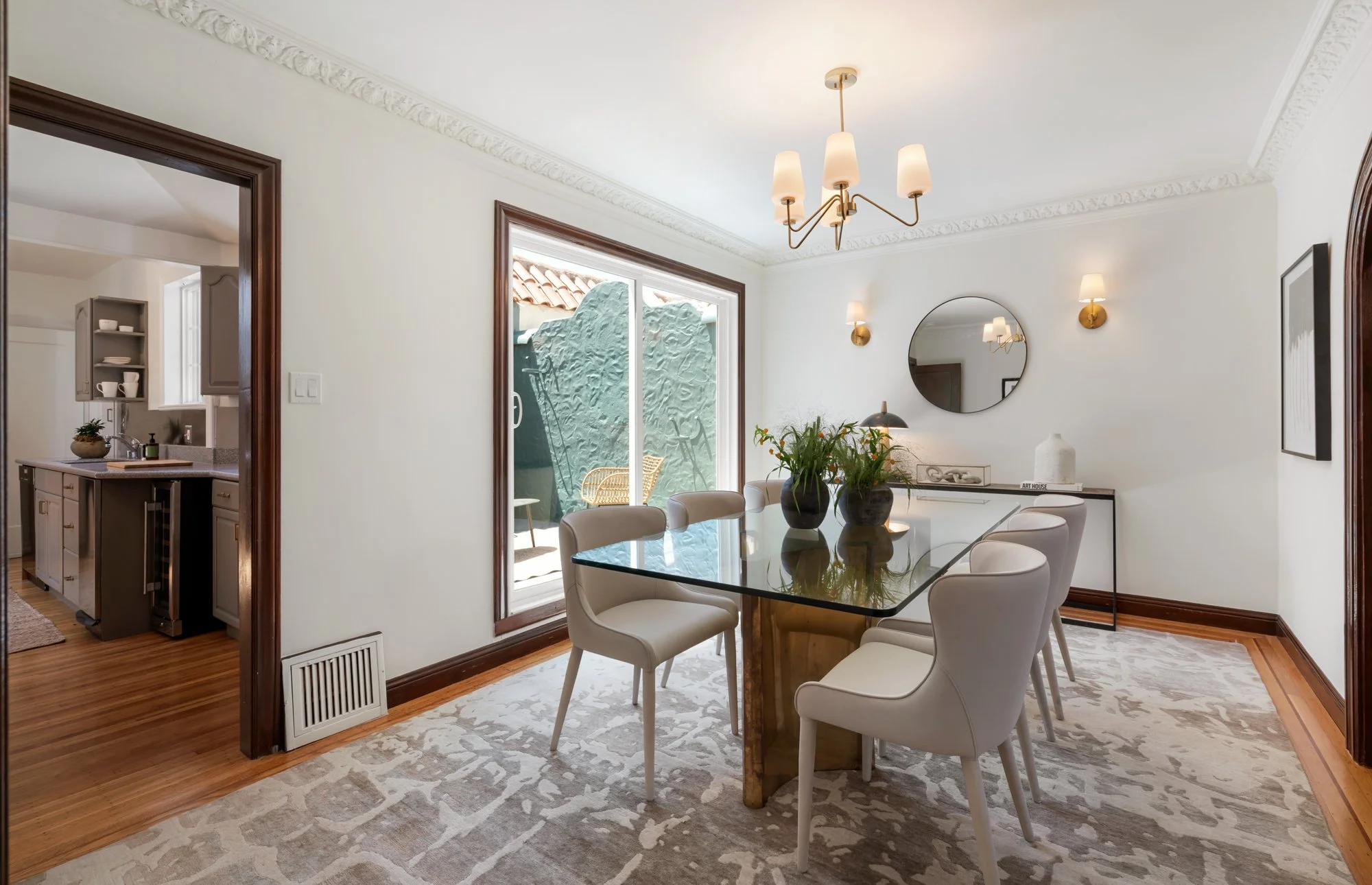 Dining room with a glass-top table, white upholstered chairs, a round mirror on the wall, plants on the table, and a sliding glass door leading to an outdoor patio. A chandelier and wall sconces provide lighting.