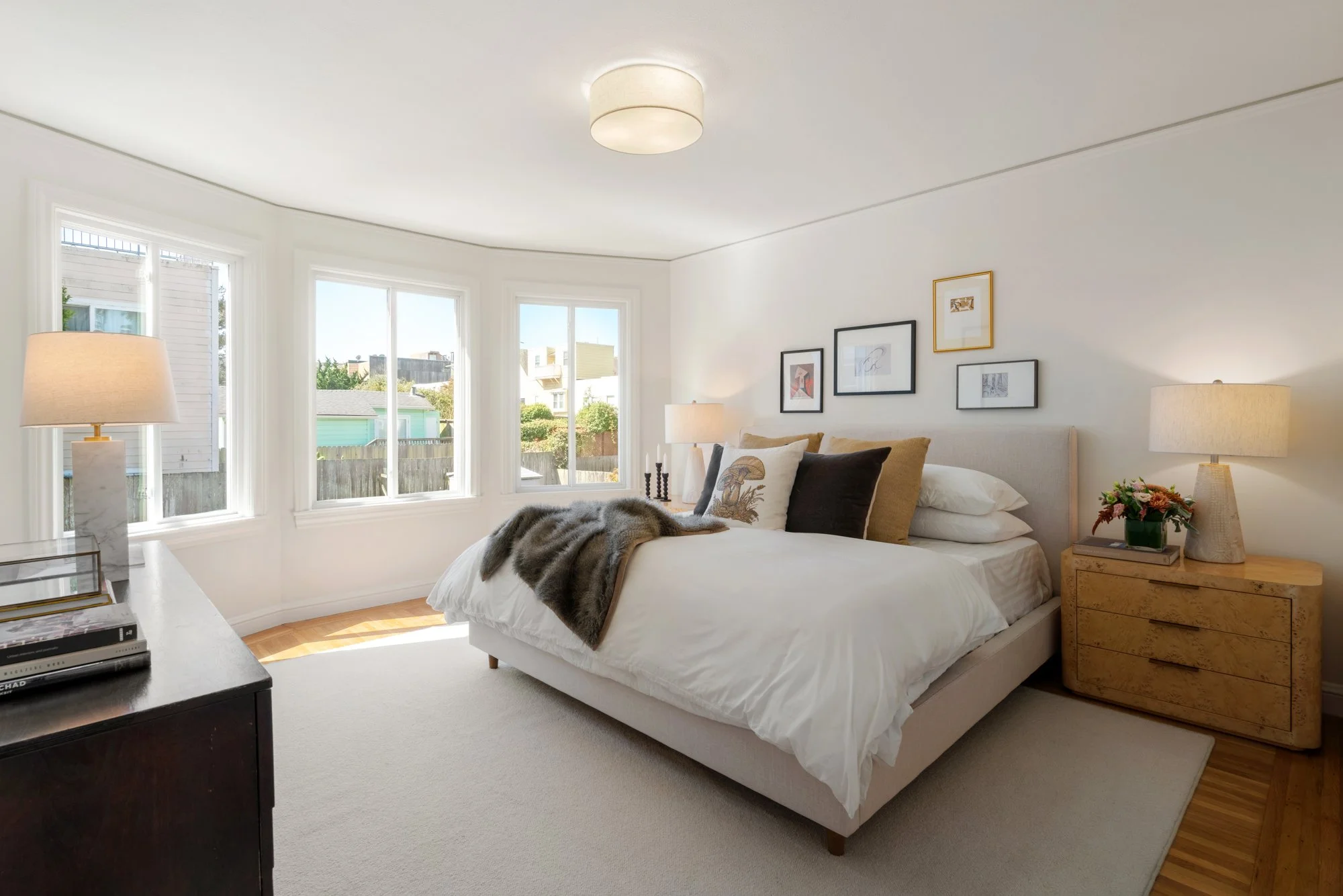 A cozy bedroom with a bed, framed pictures on the wall, lamps on bedside tables, and large windows bringing in natural light.