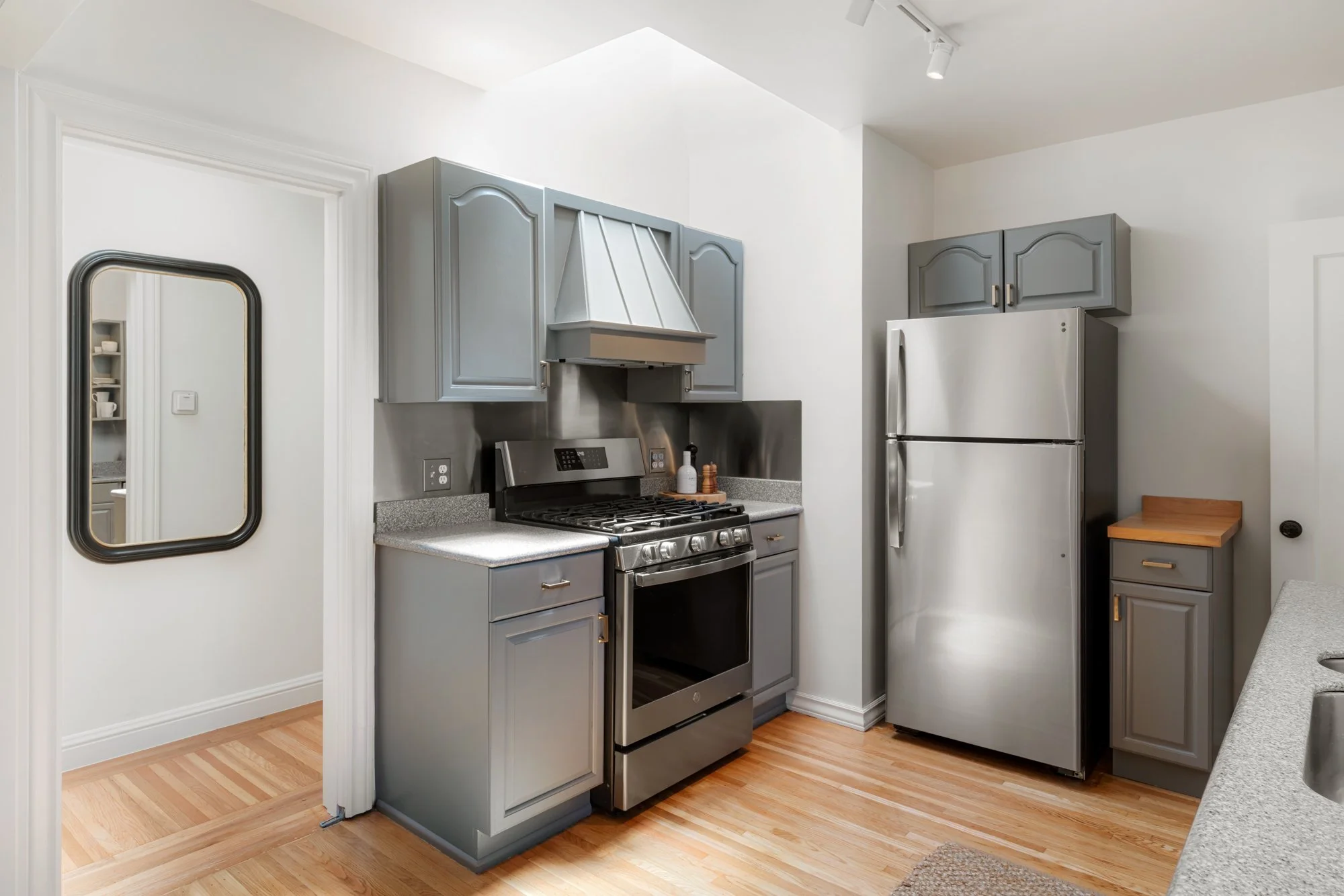 The image shows a modern kitchen with gray cabinets, a stainless steel stove, a fridge, and a small wooden drawer. There is a small mirror on the wall, and the flooring is light wood.