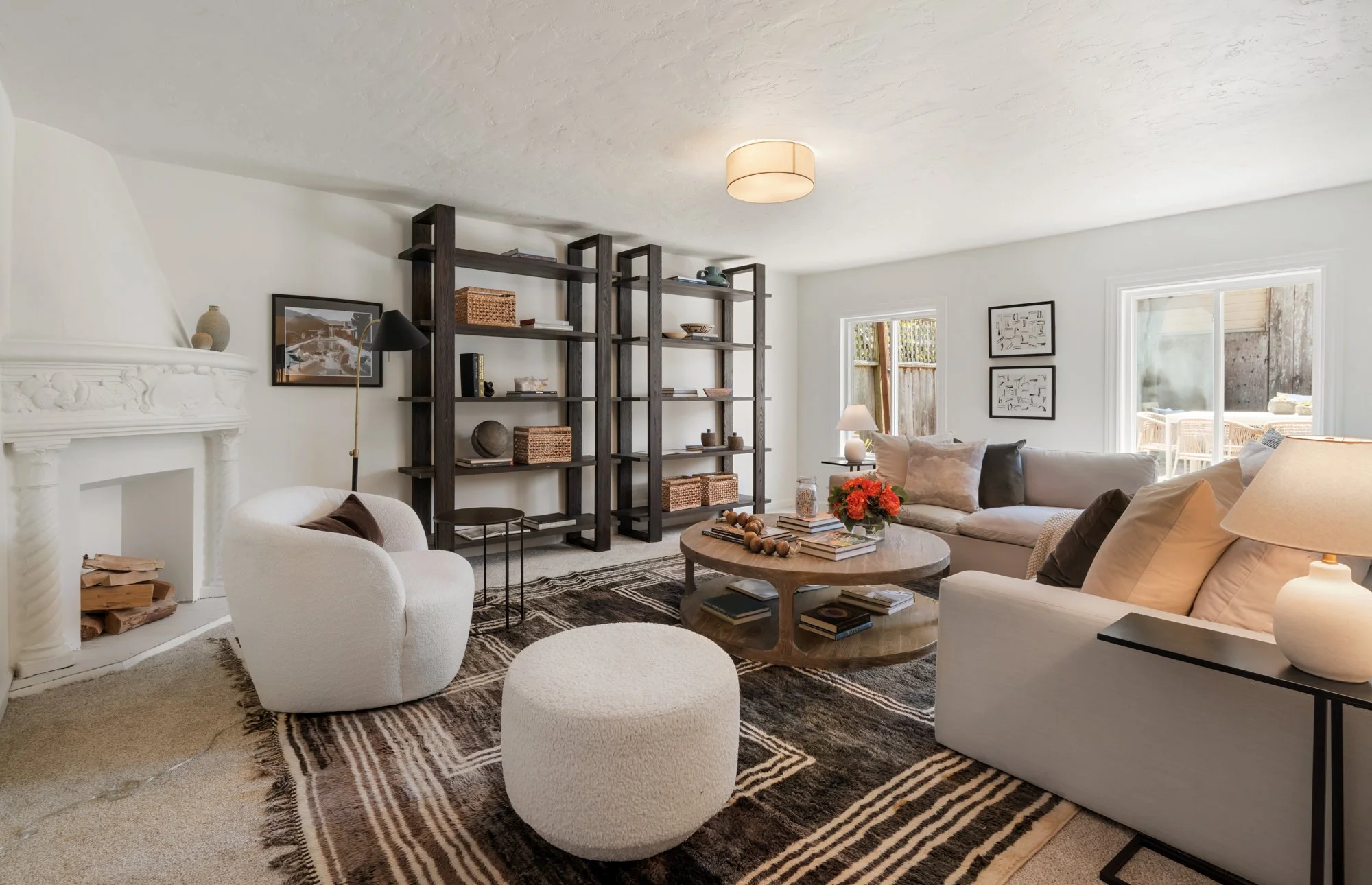 Living room with white walls and ceiling, beige carpet, and large windows. Contains white and beige sofas, black-framed artwork, a wooden coffee table with books and flowers, a tall black bookshelf with decorative items, a white armchair with a brown