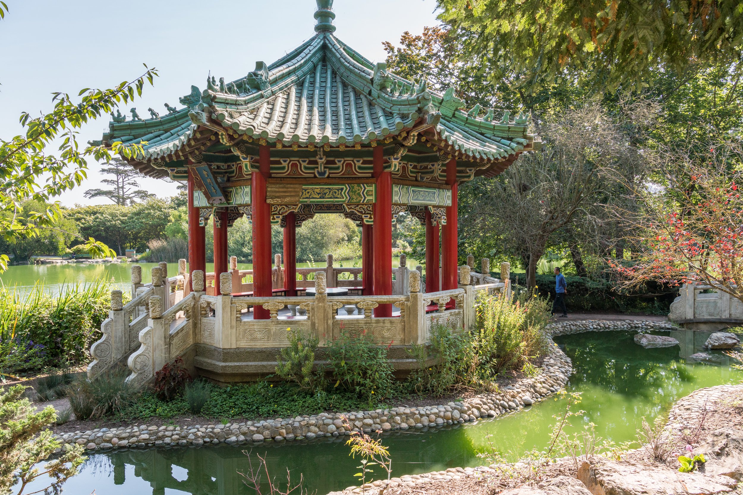 Traditional Chinese pavilion with green-tiled roof and red pillars, surrounded by lush trees and a pond in a park.