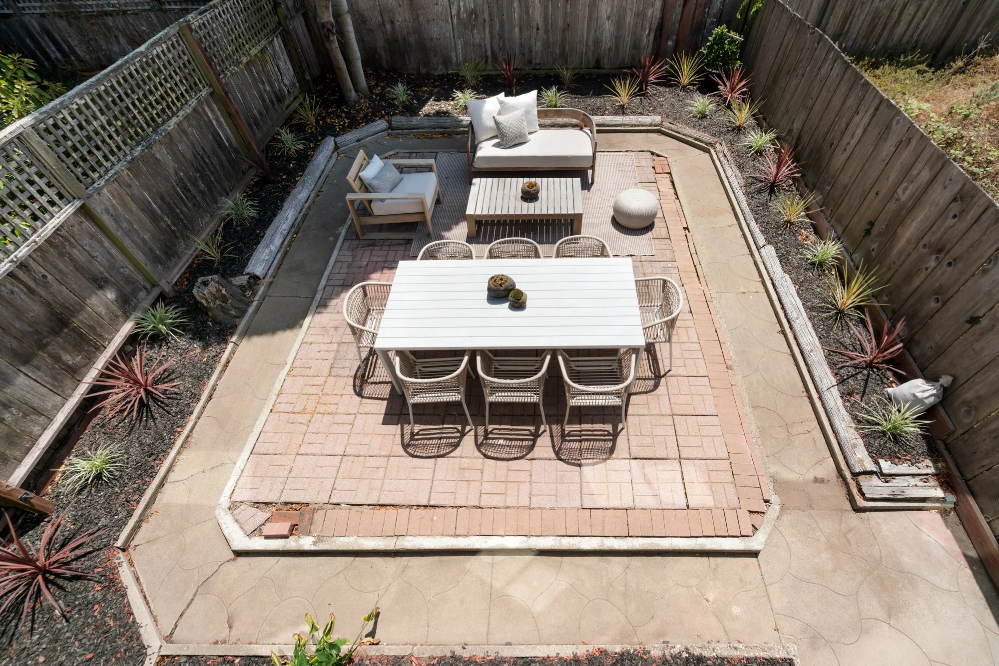 An overhead view of a small backyard patio enclosed by wooden fencing. The patio has a brick-and-concrete ground with a section of netted brick, used for outdoor dining. There is a white rectangular dining table with matching chairs, a separate sitti