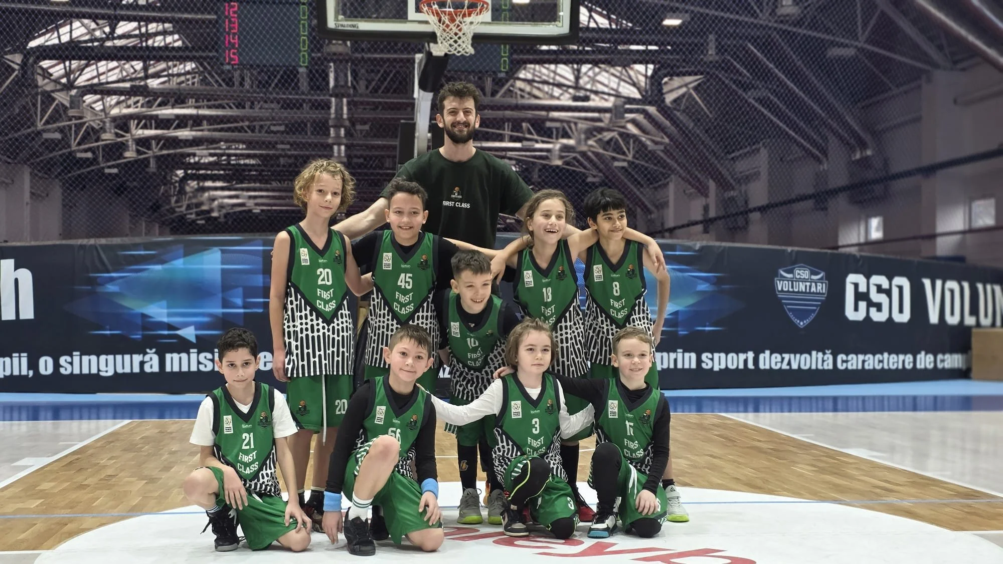 A group of young boys and girls in green basketball uniforms posing for a team photo on an indoor basketball court, with an adult coach standing behind them. The court has a basketball hoop and a digital scoreboard in the background.