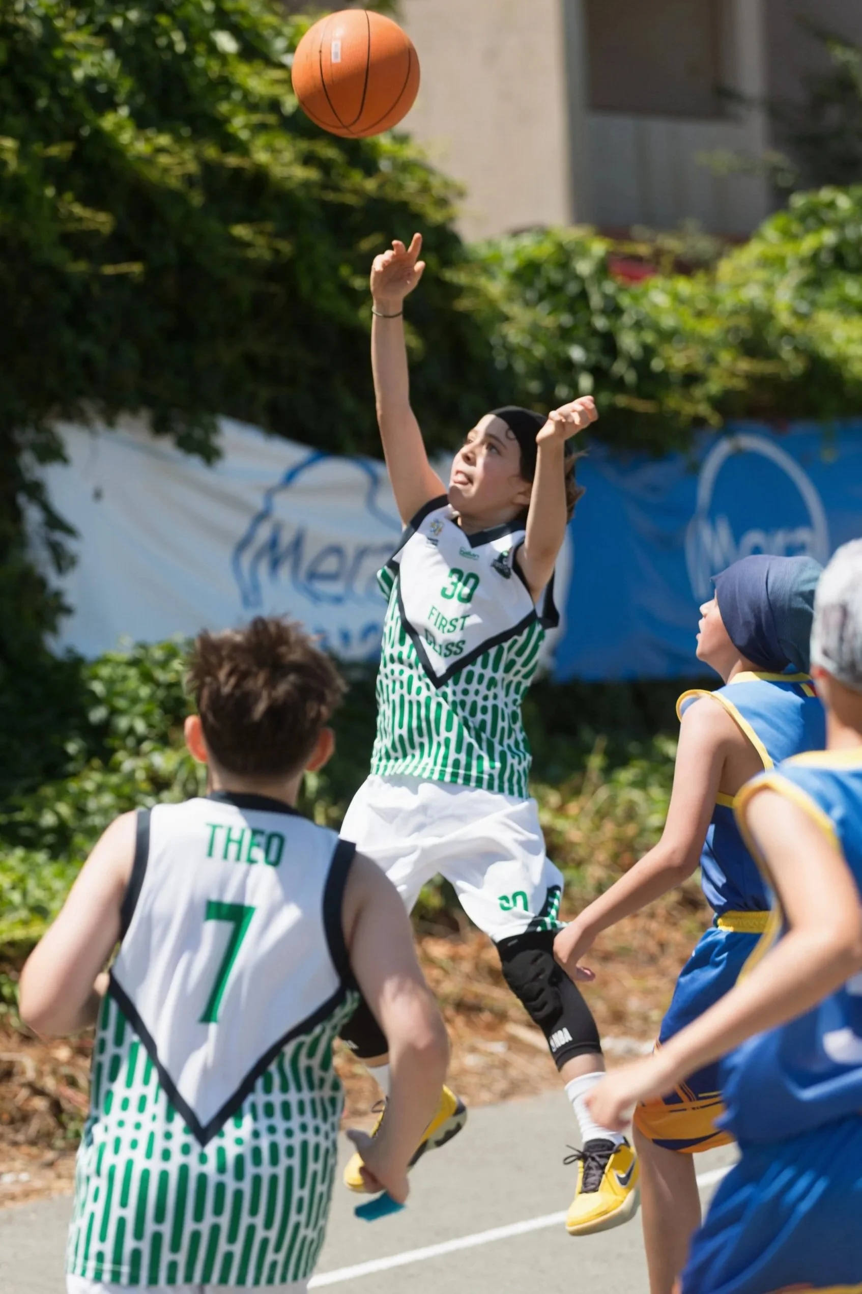 Children playing basketball outdoors, jumping to reach the basketball in the air. Others are watching and supporting. Bright daytime with greenery in the background.