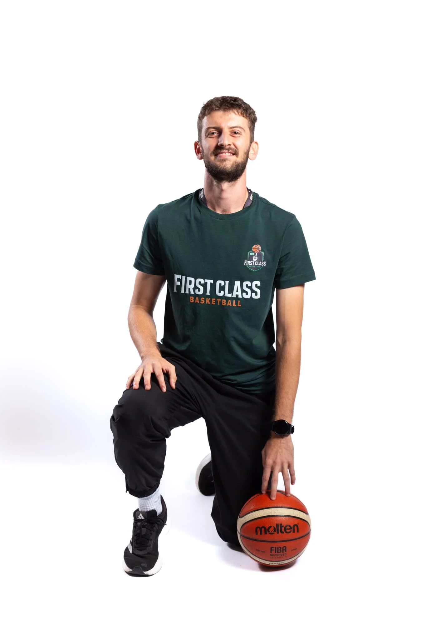 Young man in black athletic clothes, kneeling on one knee with a basketball on the floor near his hand against a white background.