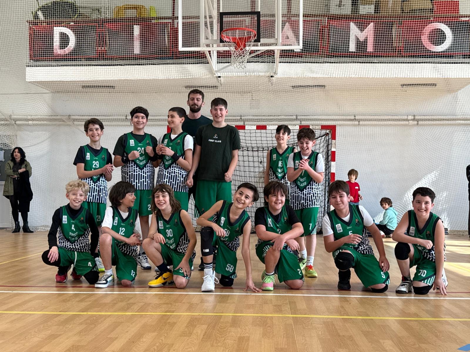 A group of young boys and their coach in a gymnasium, posing for a photo after playing basketball. The boys are wearing green and black sports uniforms, some are kneeling in the front row, others standing in the back, with smiles on their faces.