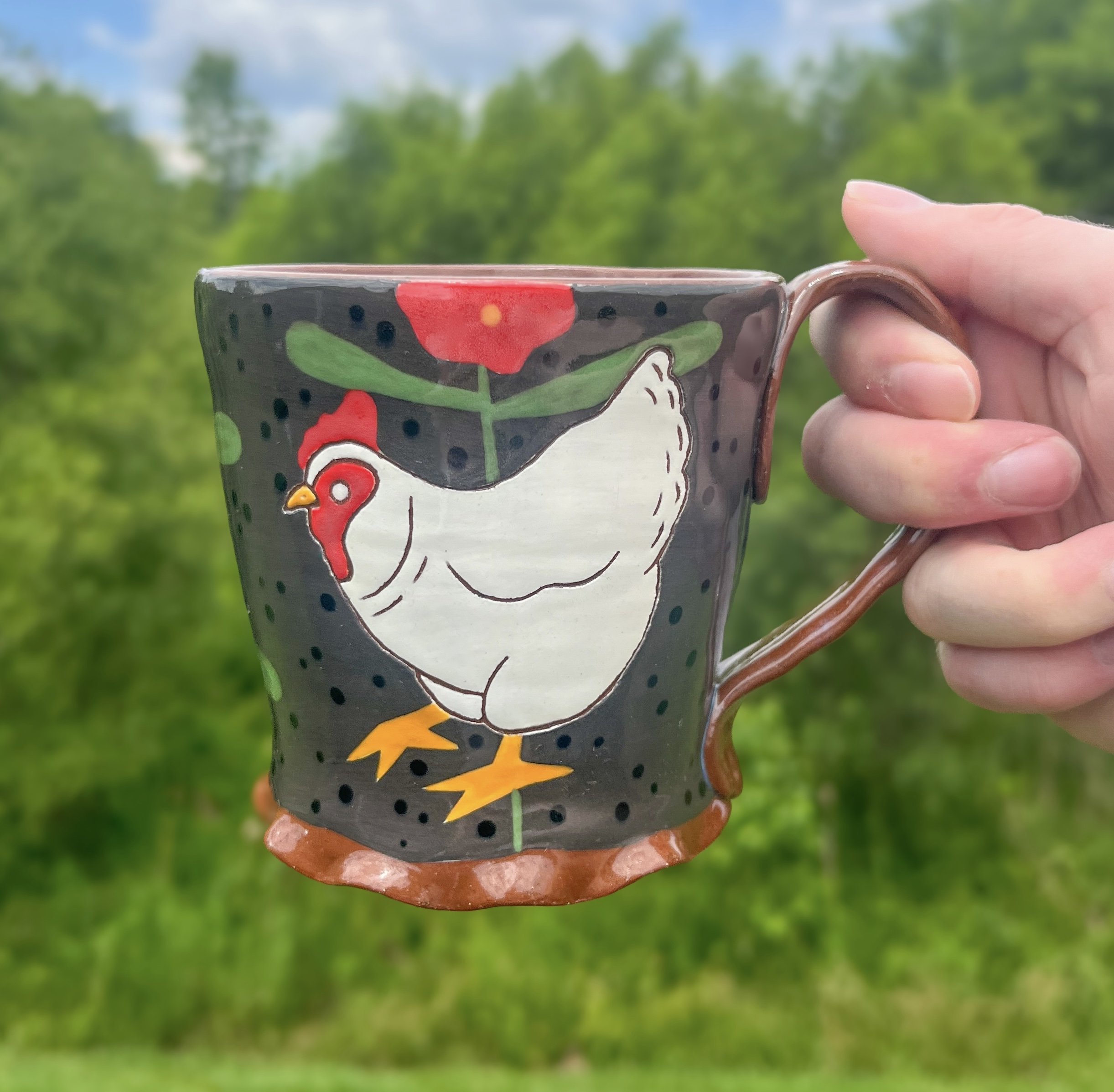 A hand holding a decorated ceramic mug with a chicken design, against a blurred green outdoor background.