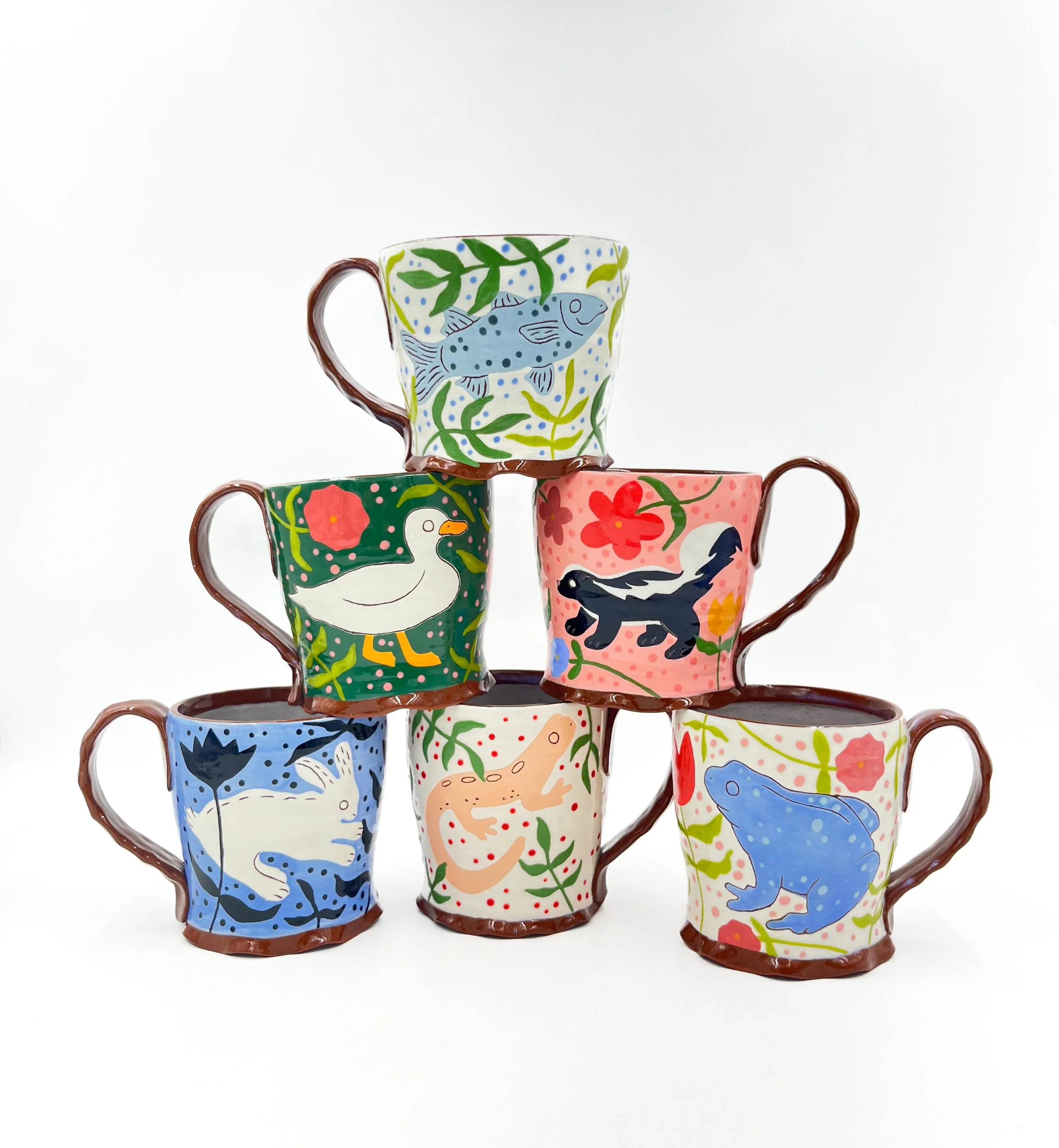 Six colorful ceramic cups decorated with animals, plants, and nature scenes, stacked in a pyramid formation against a white background.