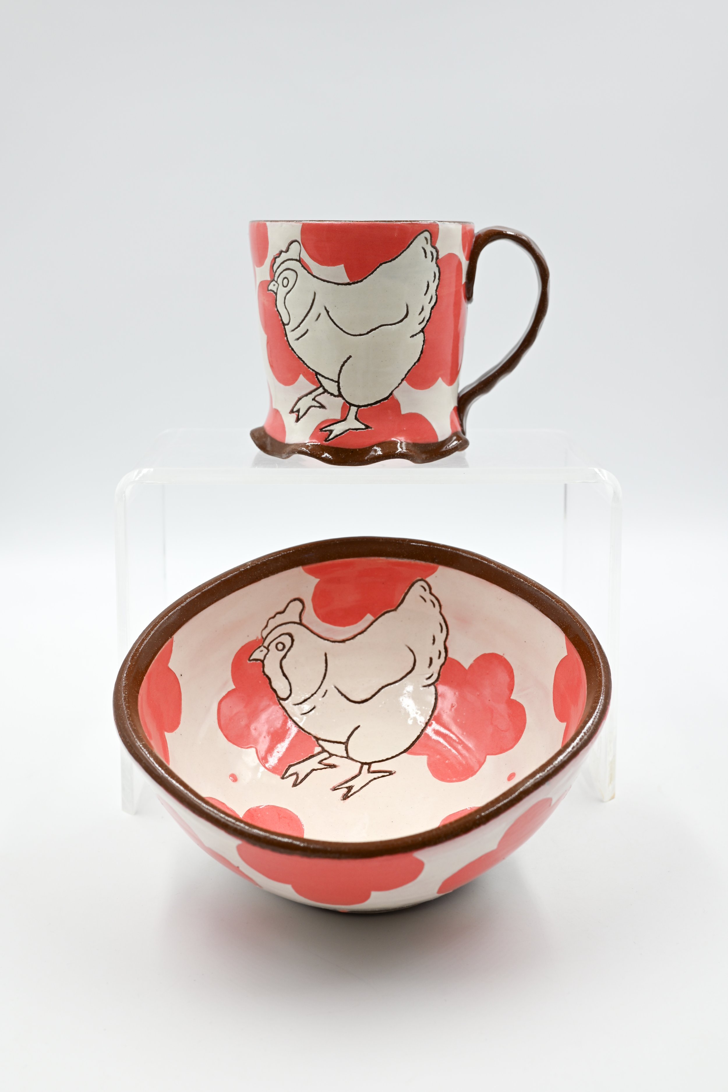 A set of ceramic dishware includes a mug, a bowl, and a plate, all decorated with a cartoon chicken and red floral patterns, displayed on a white background.