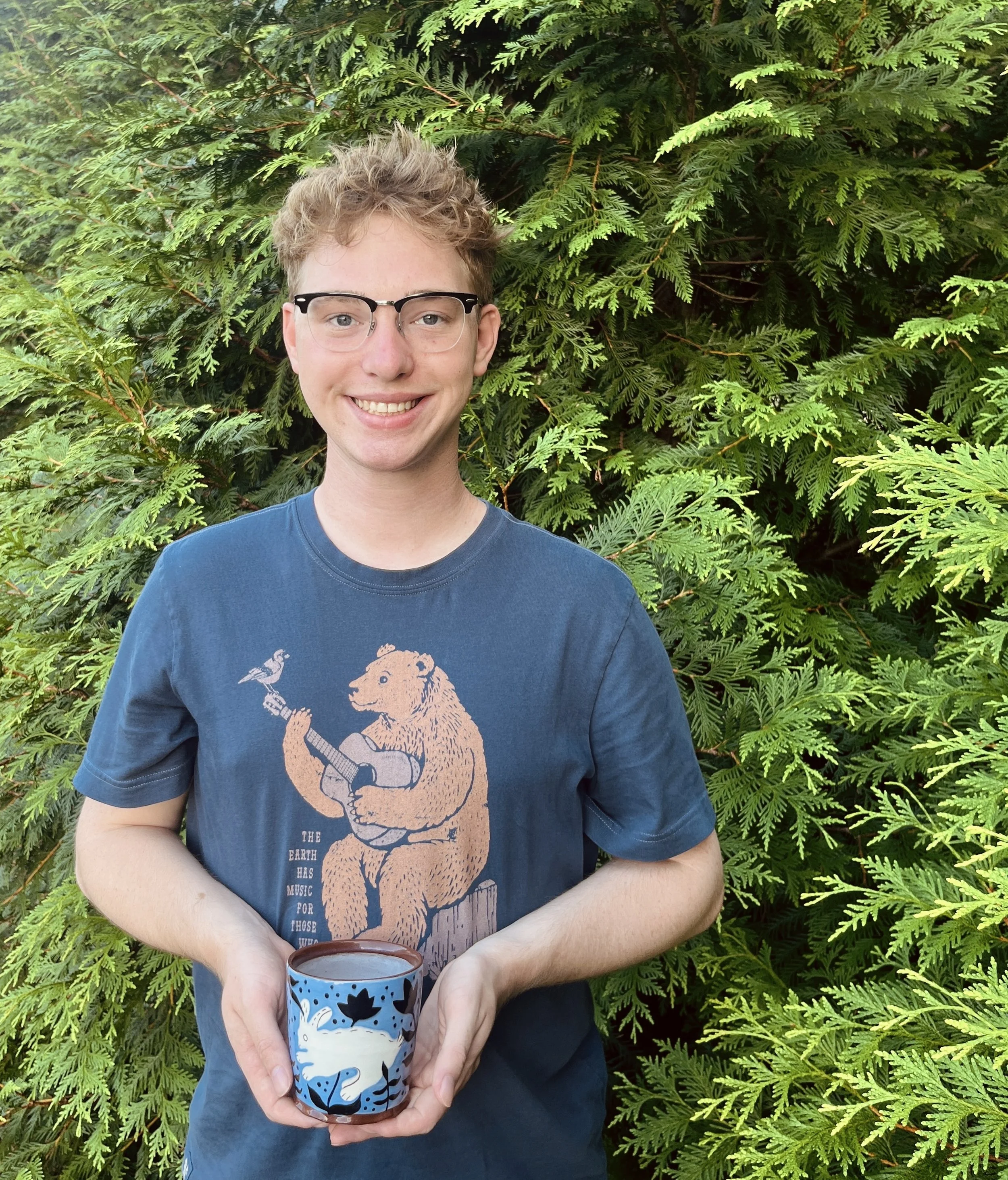 Ryan Catalano smiling while holding a patterned ceramic cup, standing in front of lush green foliage.
