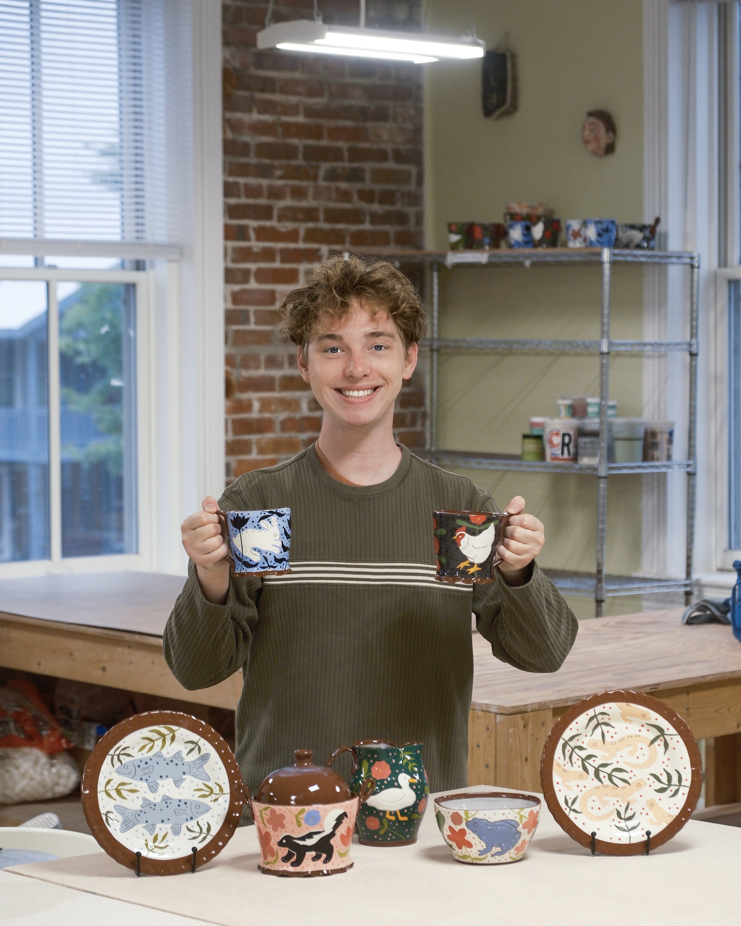 Ryan smiling and holding two decorated mugs, standing behind a display of colorful ceramic plates and teapots with animal and nature designs on a table. The background shows a brick wall, shelves with boxes, and windows.
