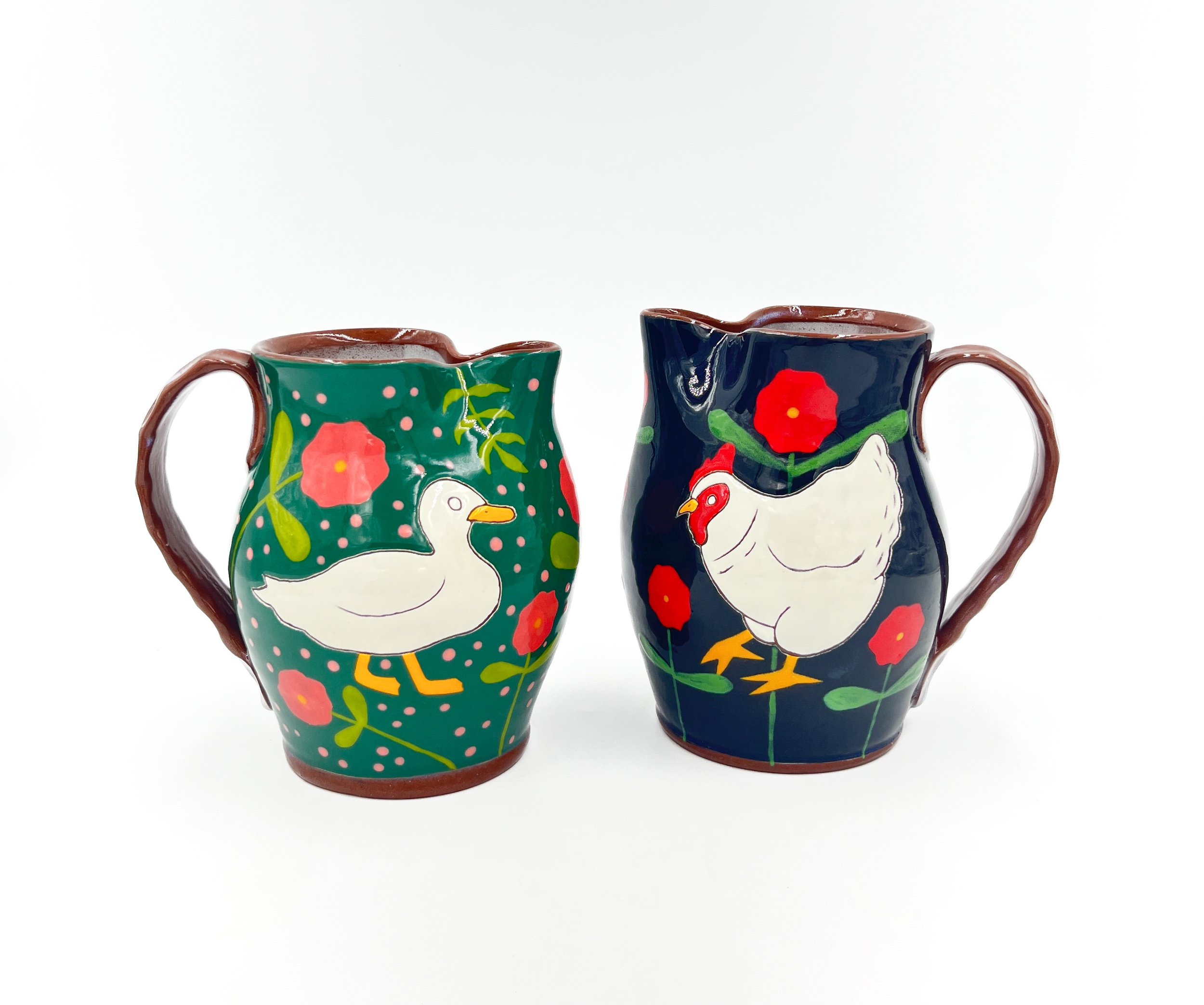 Two colorful ceramic mugs with painted rooster and floral designs, set against a plain white background.